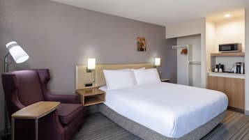 Hotelkamer met een bed, een stoel, een bureau, een nachtkastje, een lamp, een kunstwerk aan de muur, en een kitchenette met een magnetron.