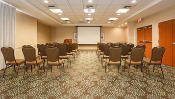 A conference room with rows of chairs facing a projection screen, a carpeted floor, and wooden doors.