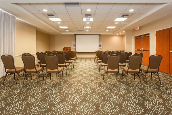 Un salón de conferencias con filas de sillas orientadas hacia una pantalla de proyección, suelo alfombrado y puertas de madera.