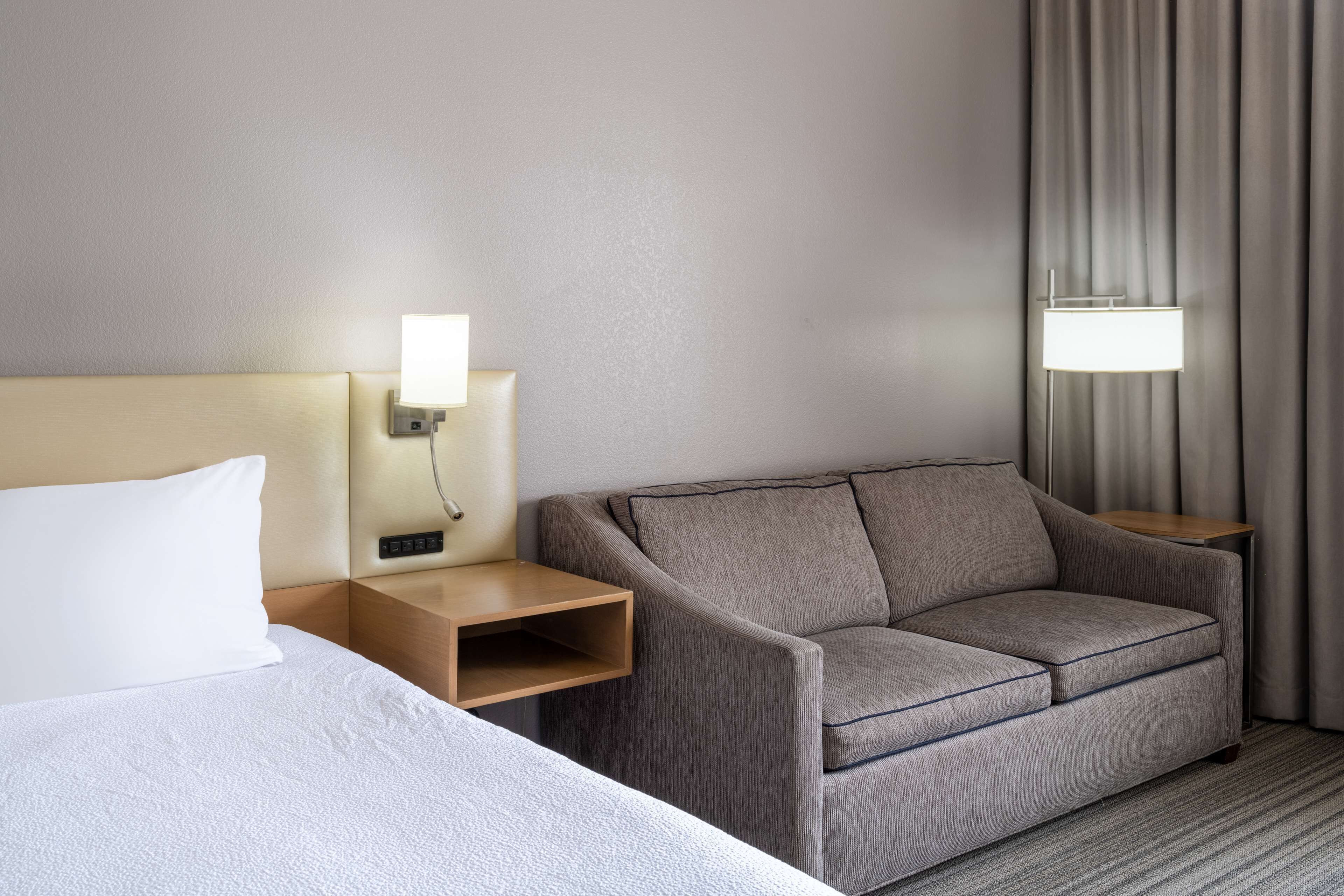 A hotel room with a bed, a grey sofa, a nightstand with a lamp, and a view of curtains.