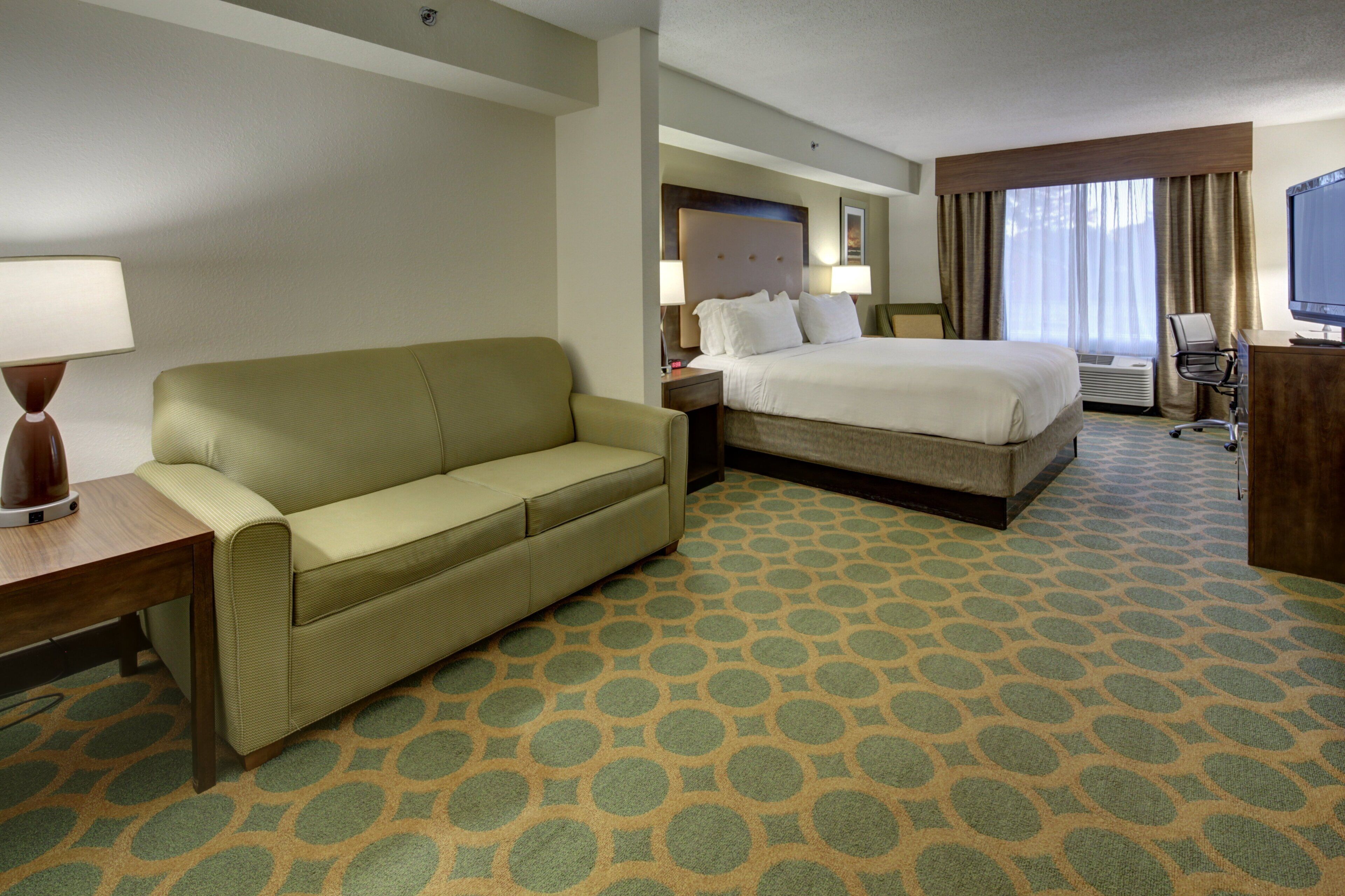 A hotel room with a bed, a sofa, a desk with a lamp, a TV, and a window with curtains.