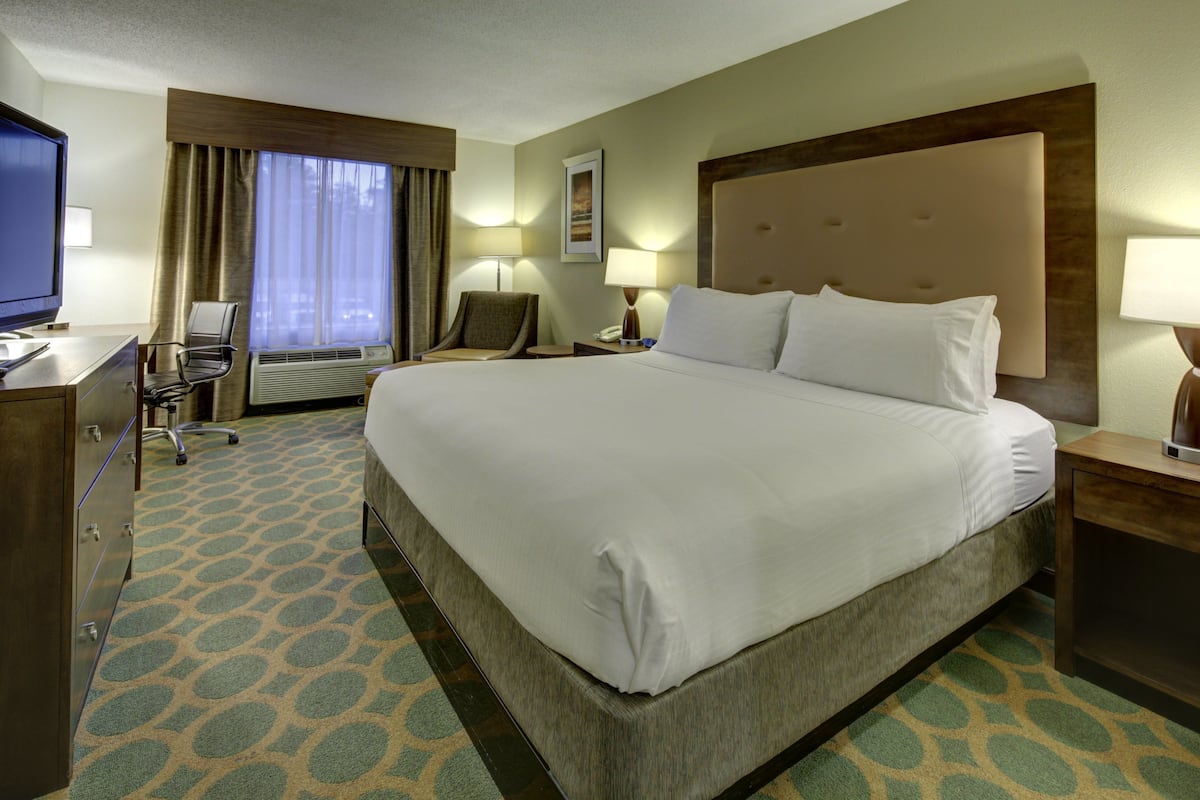 A hotel room with a large bed, a desk with a computer, a chair, a desk lamp, and a window with curtains.