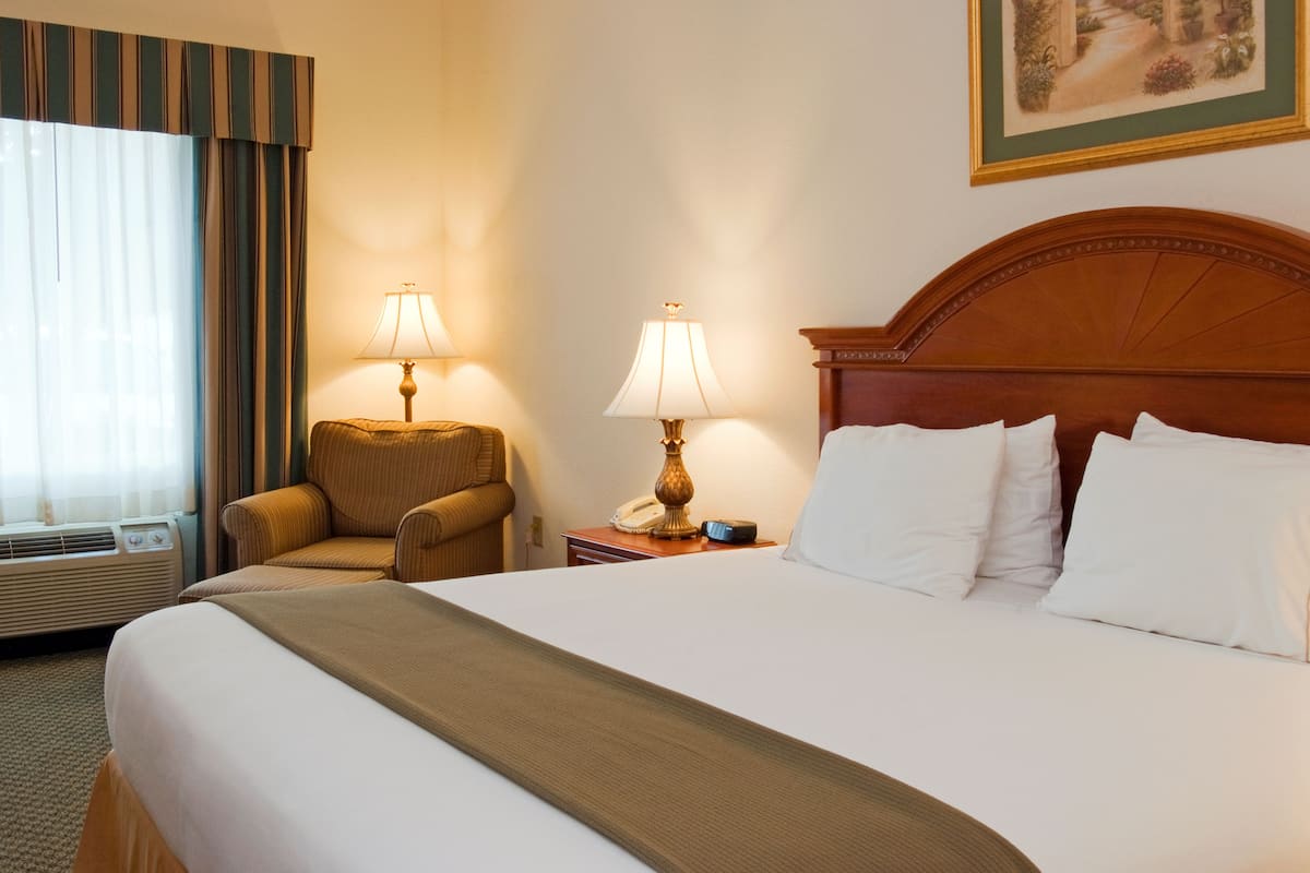 A hotel room with a large bed, two bedside lamps, a chair, a bedside table with a phone, and a window with curtains.