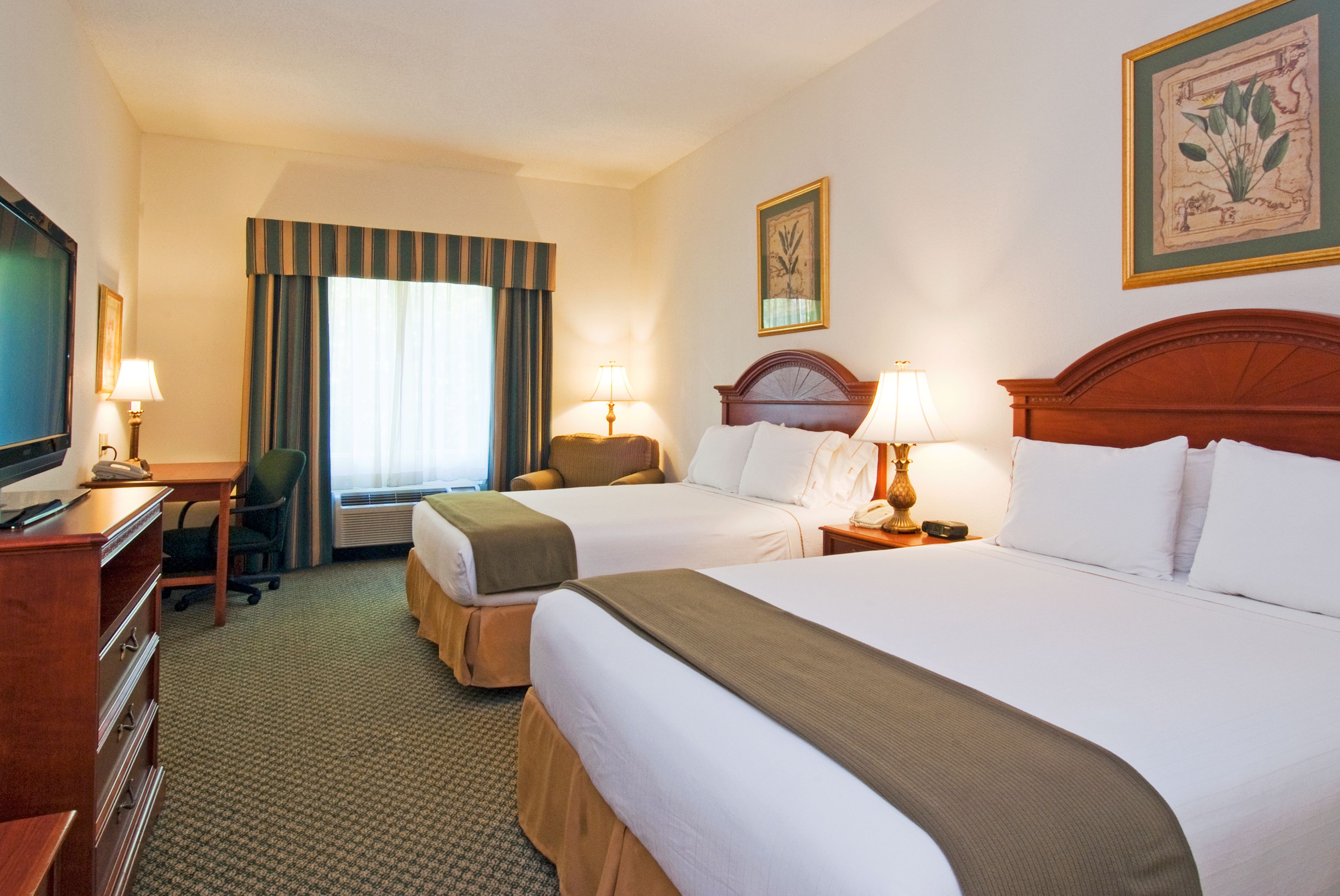 A hotel room with two beds, a desk with a chair, a television, and a large window with curtains.