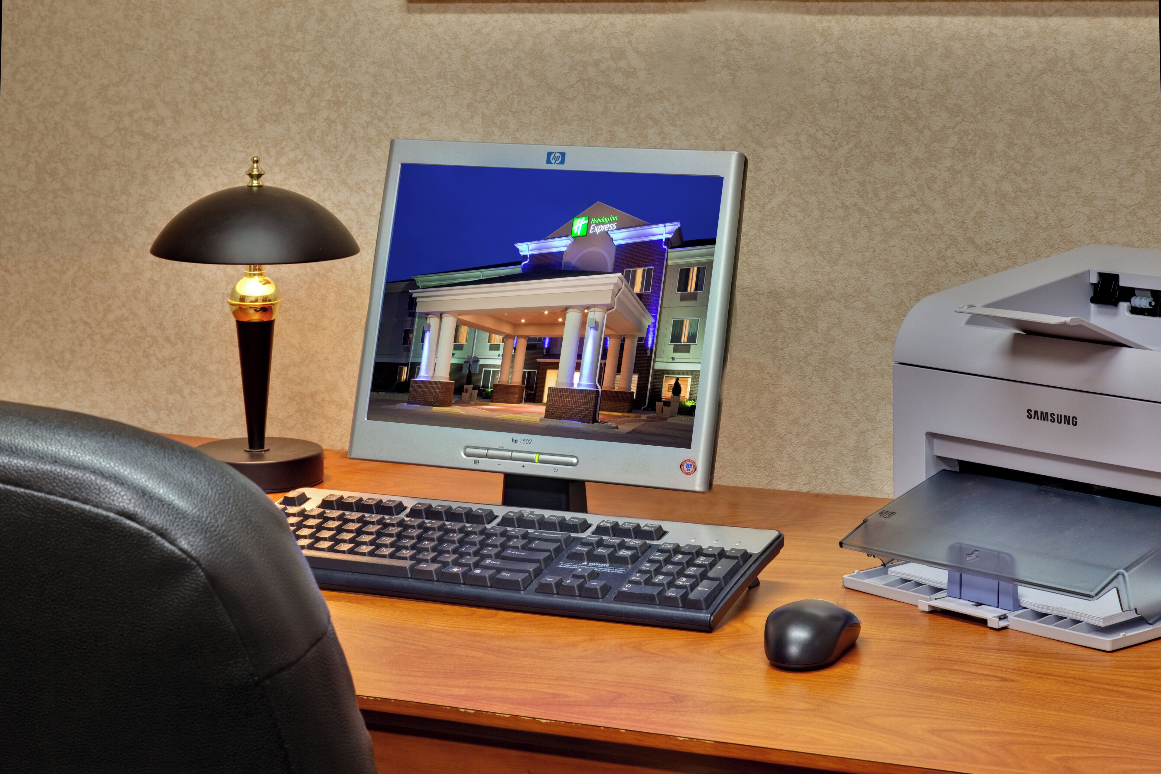 A desktop computer with a monitor displaying a hotel exterior, a keyboard, a mouse, and a printer on a desk.