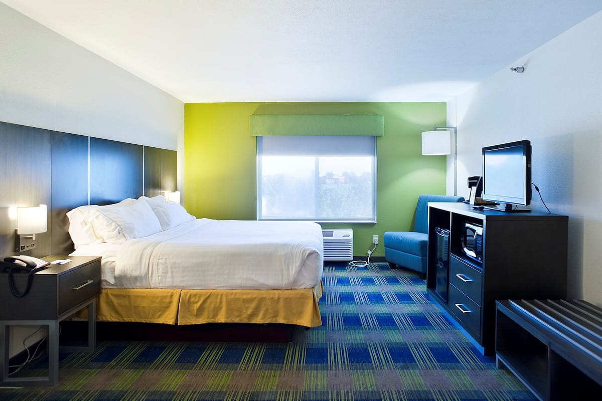 A hotel room with a bed, a desk with a lamp, a television, and a window with a view of trees.