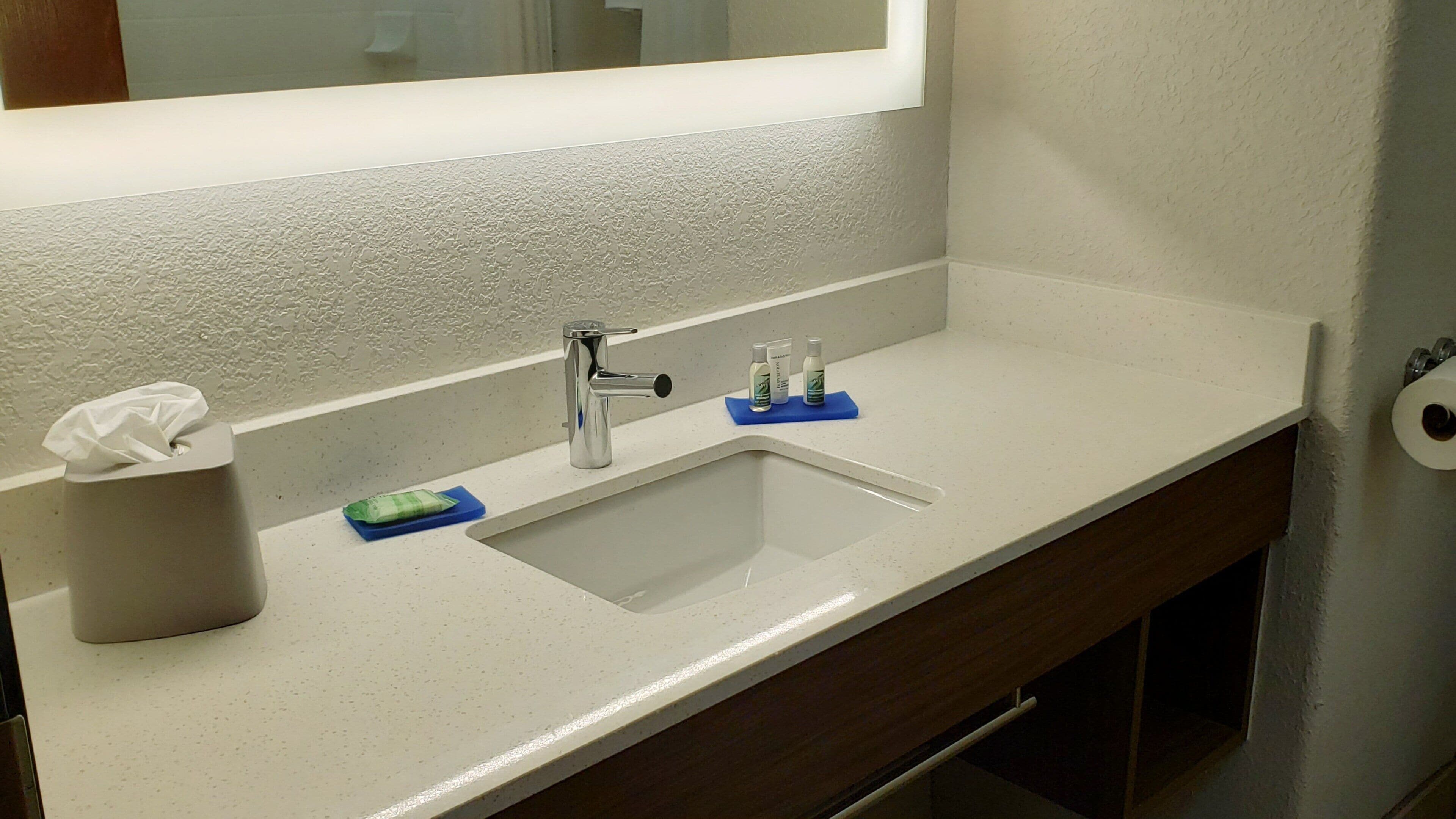 A bathroom with a white sink, a mirror, a tissue roll, and a soap dispenser.