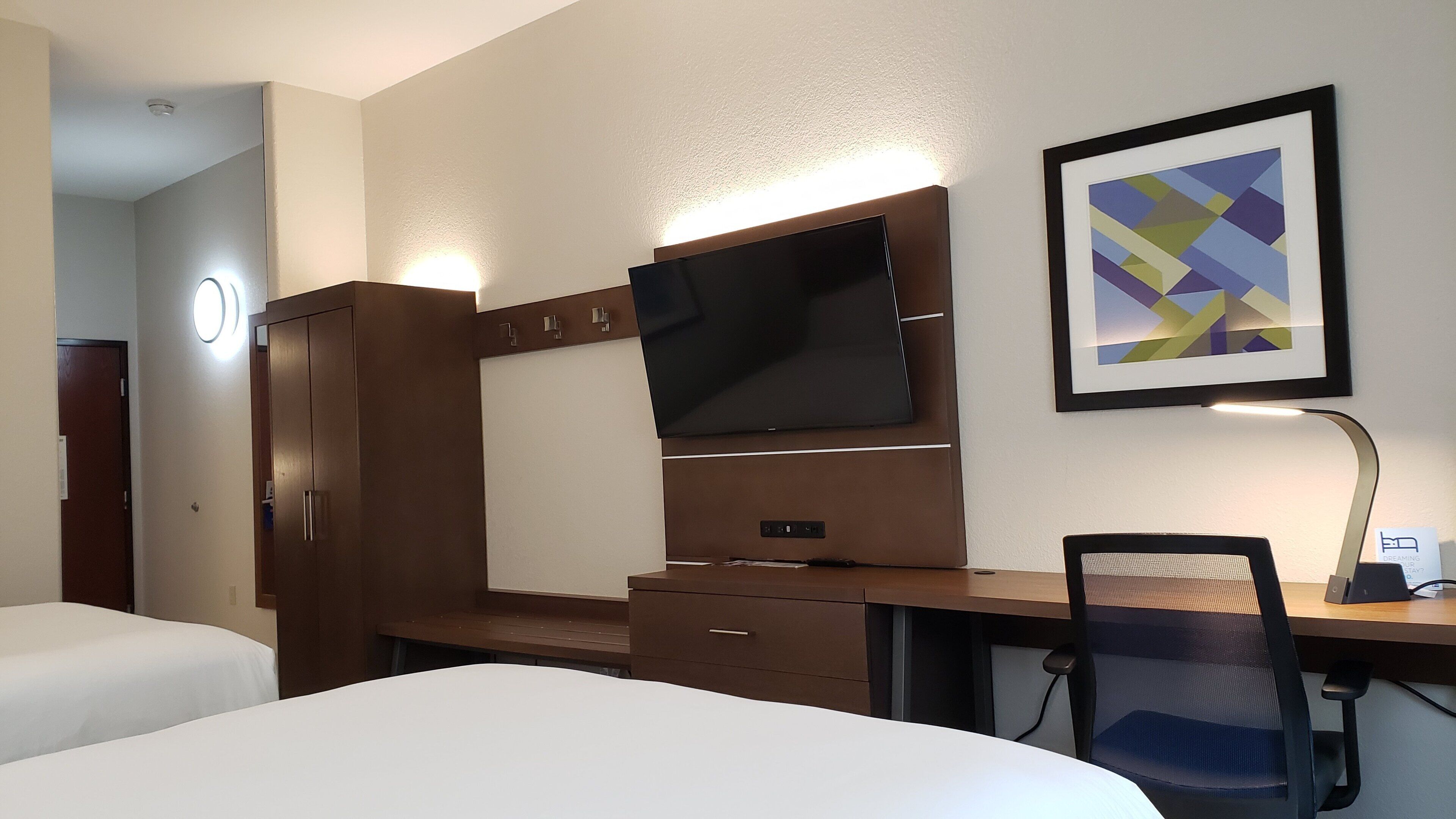 Een hotelkamer met een bed, een bureau met een computer, een televisie, een stoel en een ingelijste foto aan de muur.