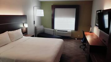 A hotel room with a bed, a chair, a desk with a TV, a lamp, and a window with blinds.