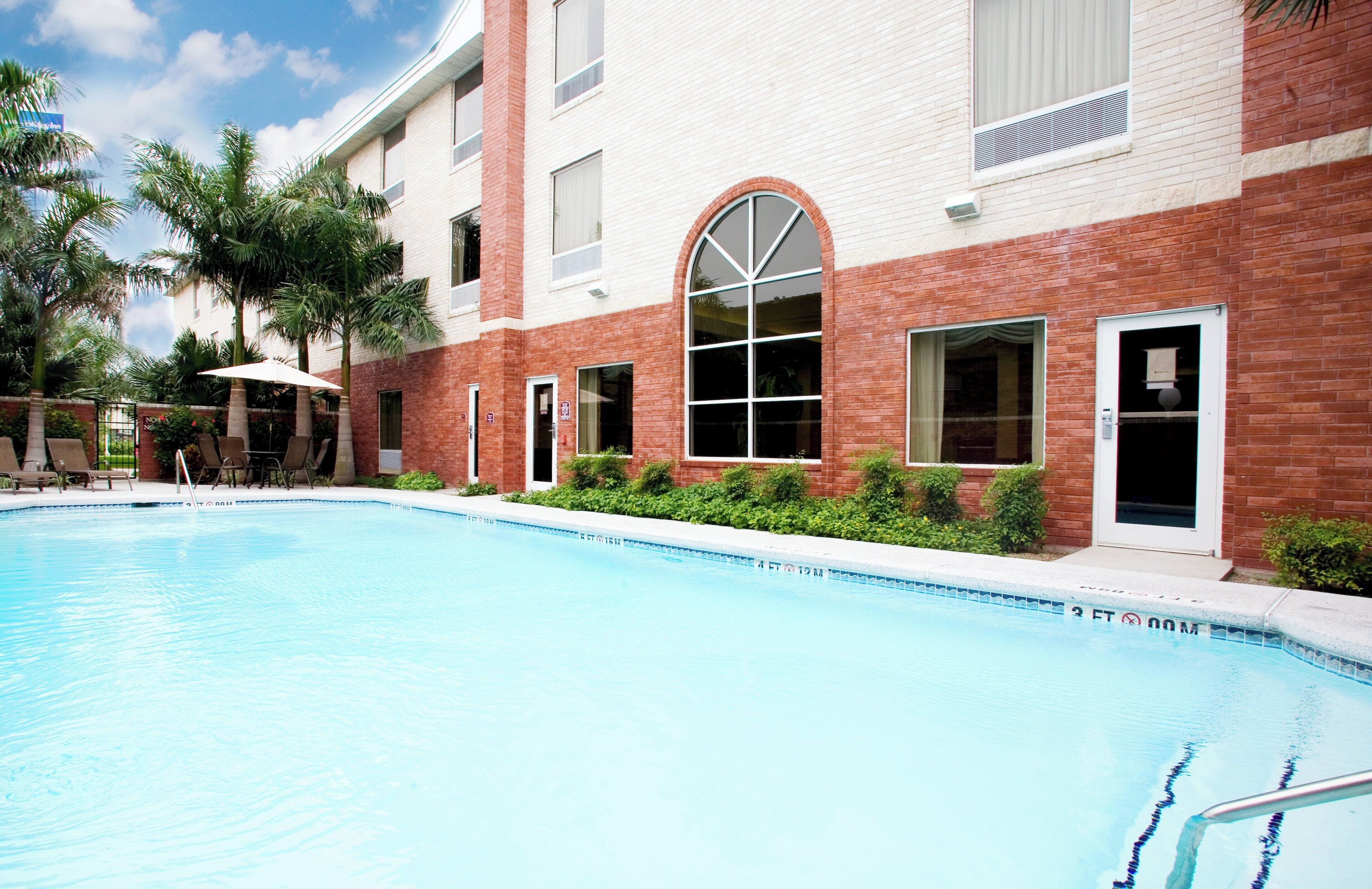 A swimming pool with lounge chairs and umbrellas, surrounded by a building with brick and white walls, and palm trees.