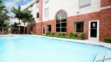 A swimming pool with lounge chairs and umbrellas, surrounded by a building with brick and white walls, and palm trees.