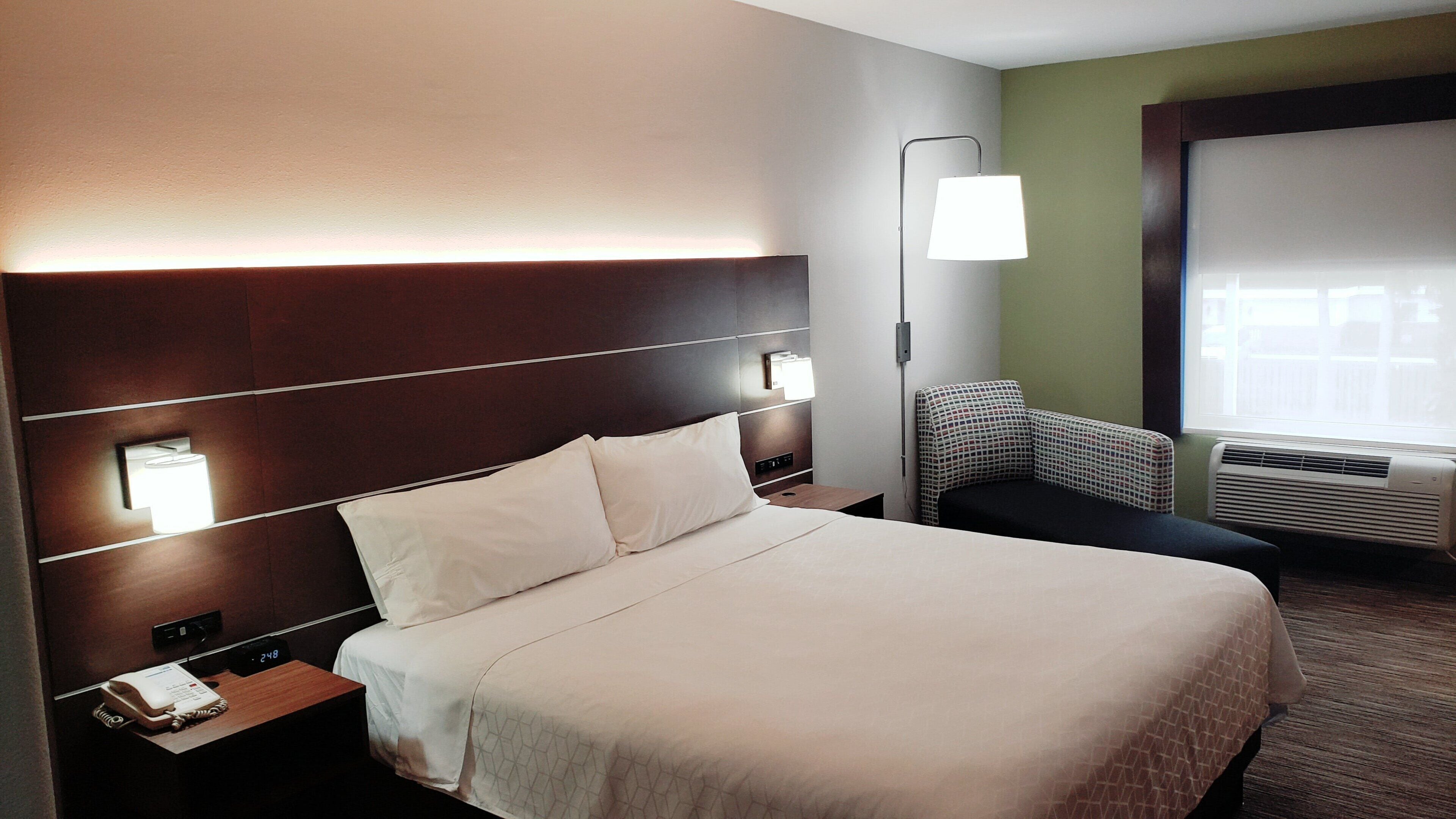 A hotel room with a large bed, bedside lamps, a chair, a bedside table with a phone, and a window with blinds.