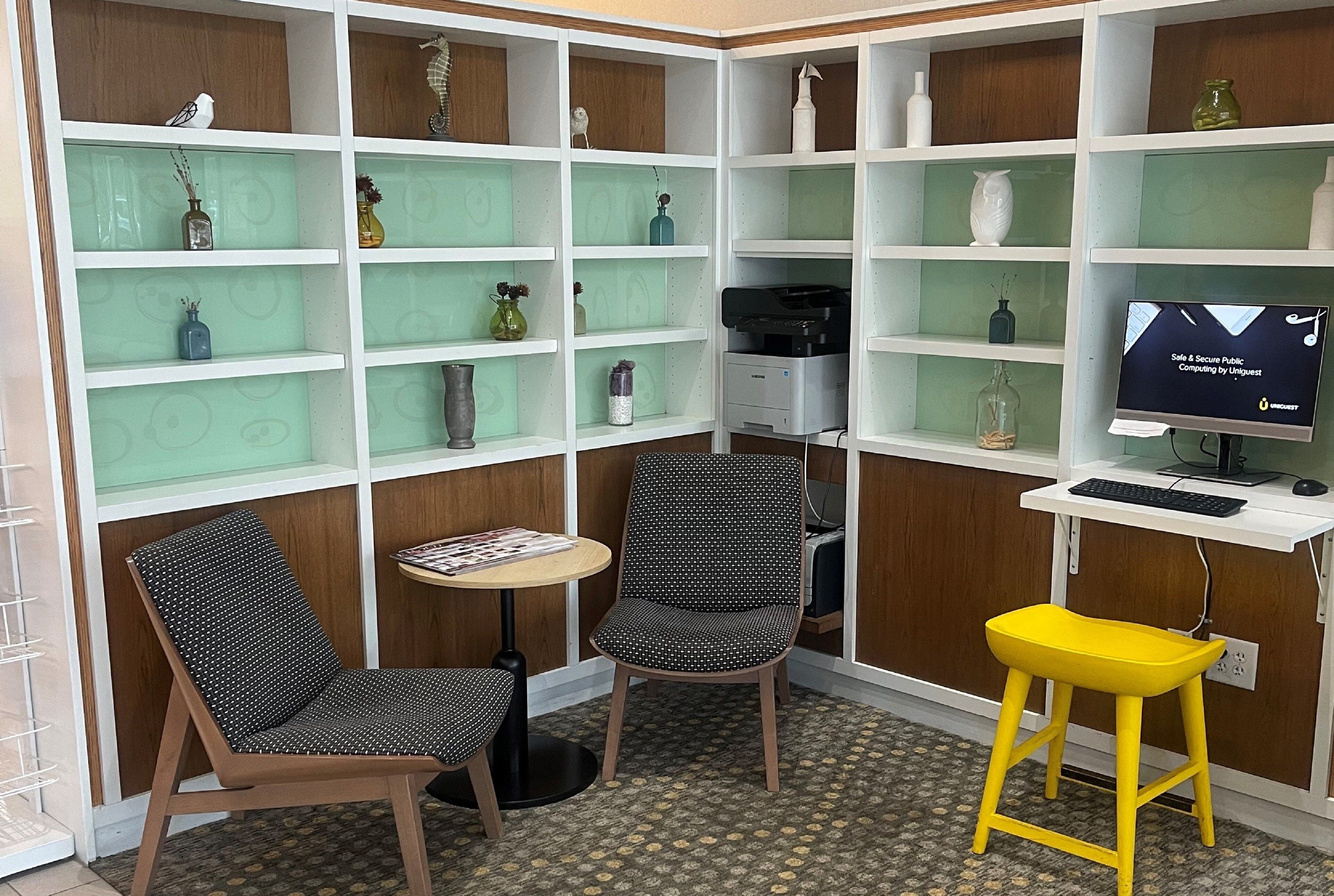 A corner office with two chairs, a small table, and a desk with a computer and printer. Shelves with decorative items line the walls.