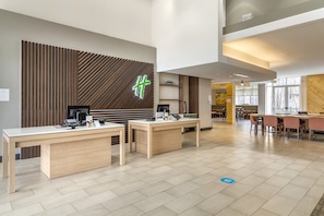 A modern reception area with wooden paneling, a green illuminated sign, and a counter with computers and a phone. - Holiday Inn Atlanta-Gas South Arena Area by IHG (Duluth)
