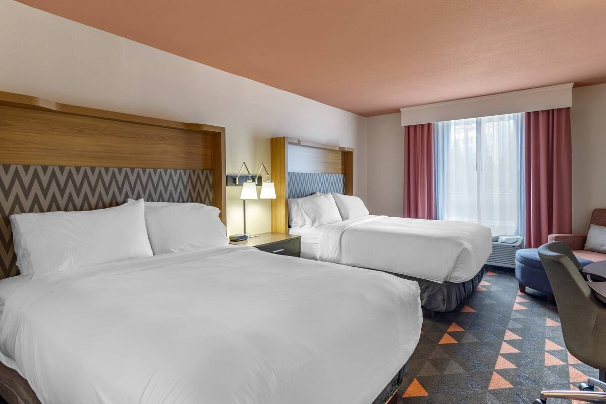 A hotel room with two beds, a desk with a lamp, a chair, and a large window with curtains.