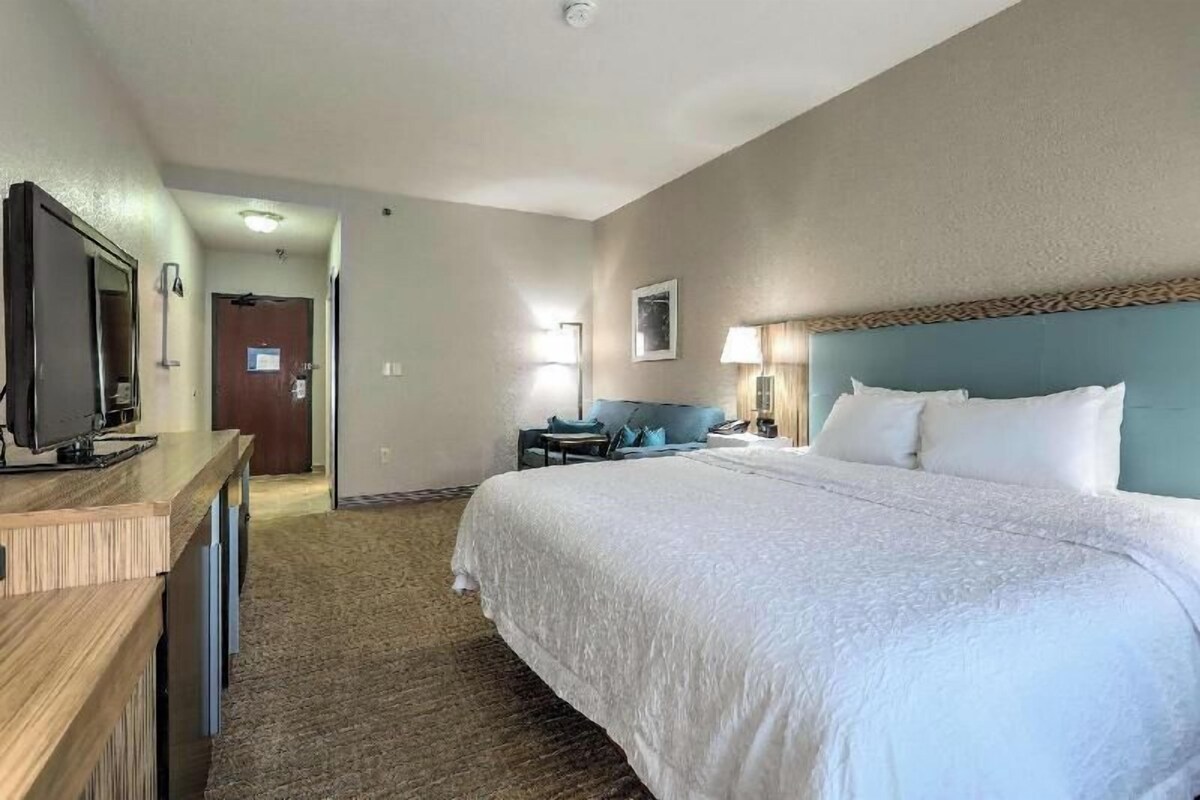 A hotel room with a large bed, a desk with a television, a chair, and a lamp.