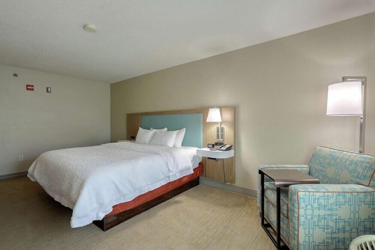 Una camera d'hotel con un letto, un comodino, una lampada e una poltrona con motivo decorativo.