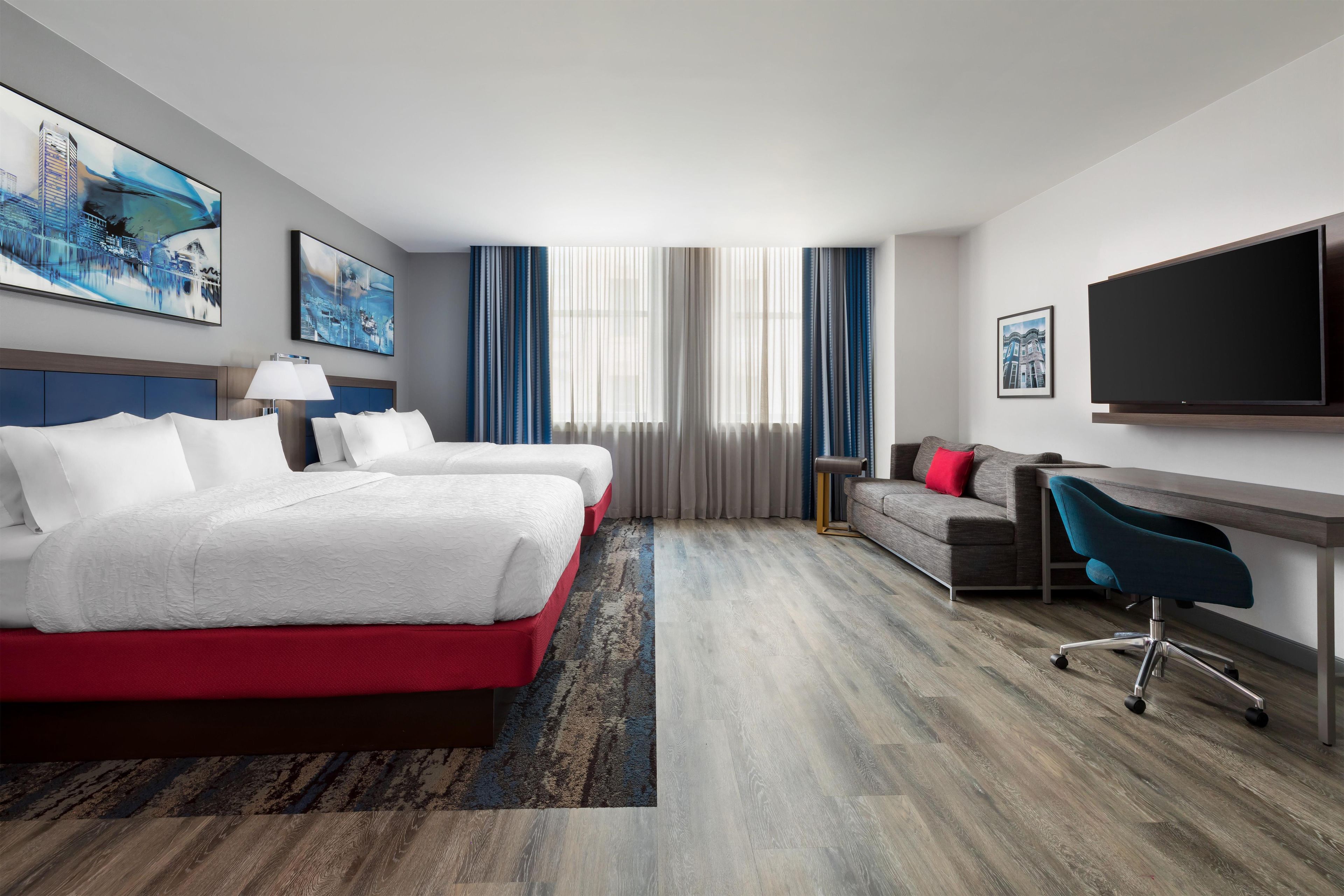 Habitación de hotel con dos camas, un escritorio, una silla, un televisor y obras de arte en las paredes.