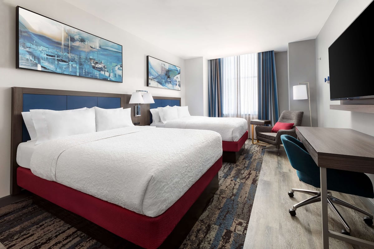 Ein Hotelzimmer mit zwei Betten, einem Schreibtisch, einem Sessel, einem Fernseher und Bildern an den Wänden.