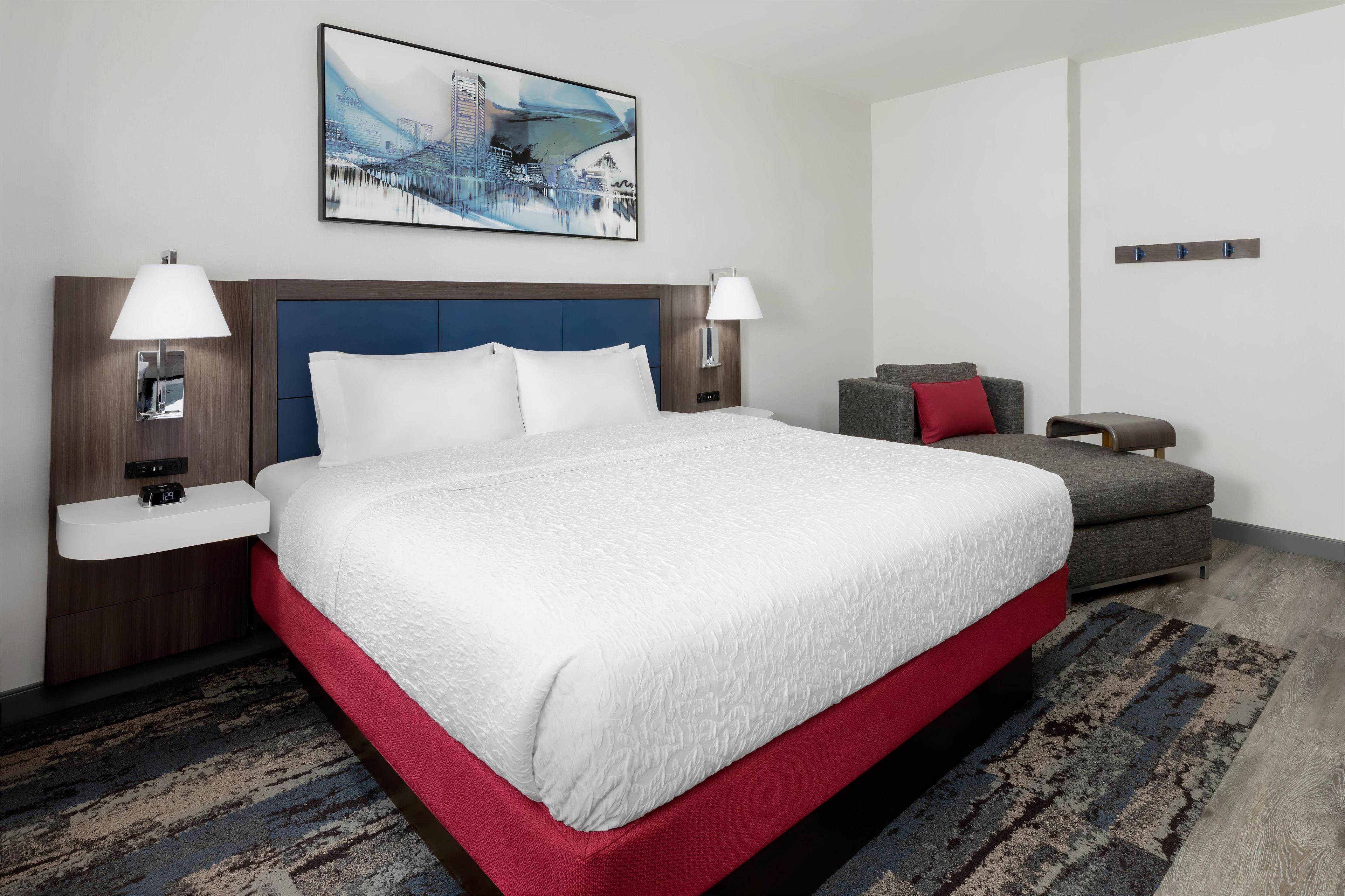 Habitación de hotel con una cama grande, dos mesitas de noche con lámparas, un sofá gris y un cojín rojo decorativo.