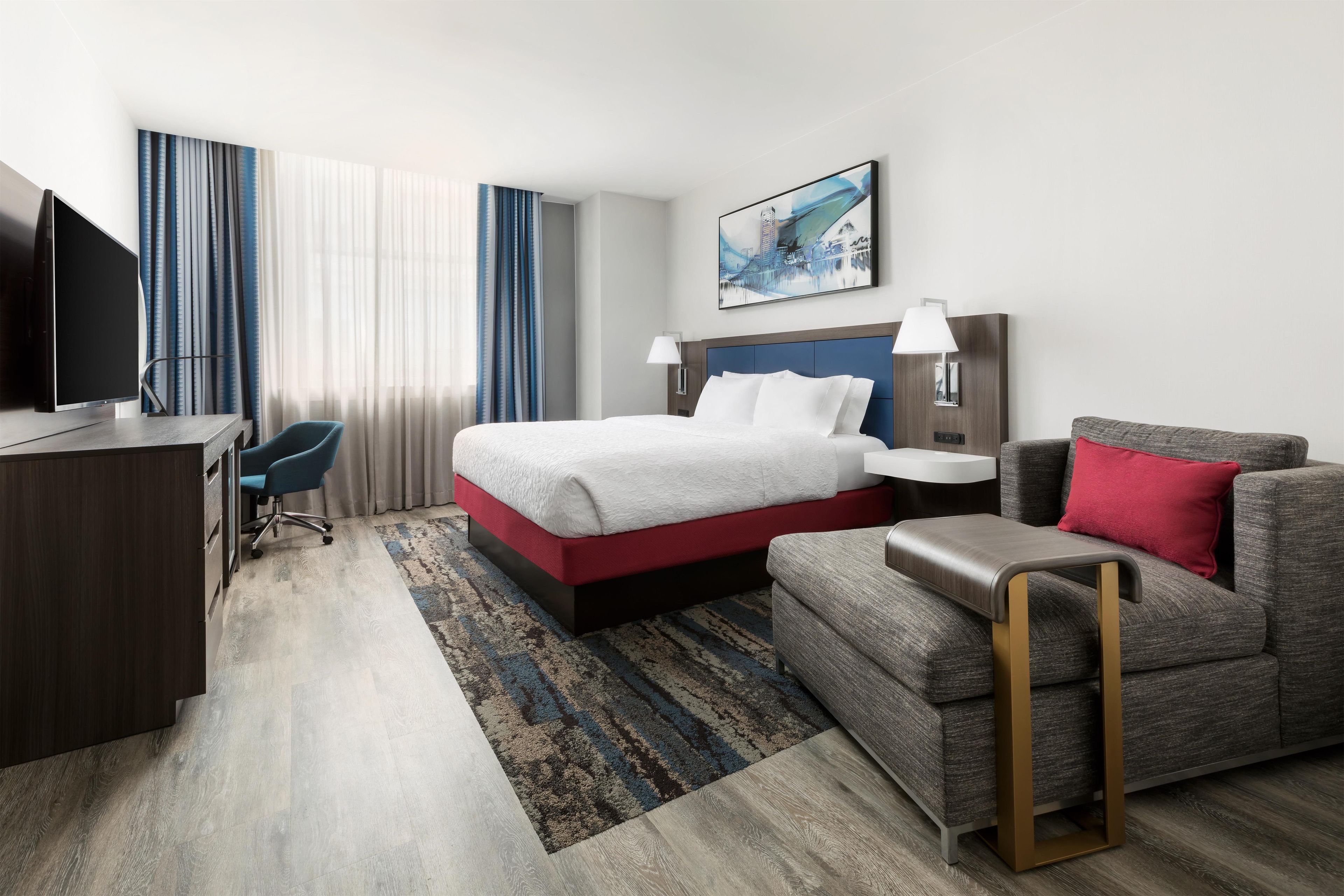 A hotel room with a bed, a desk, a TV, a chair, a sofa, and a rug.