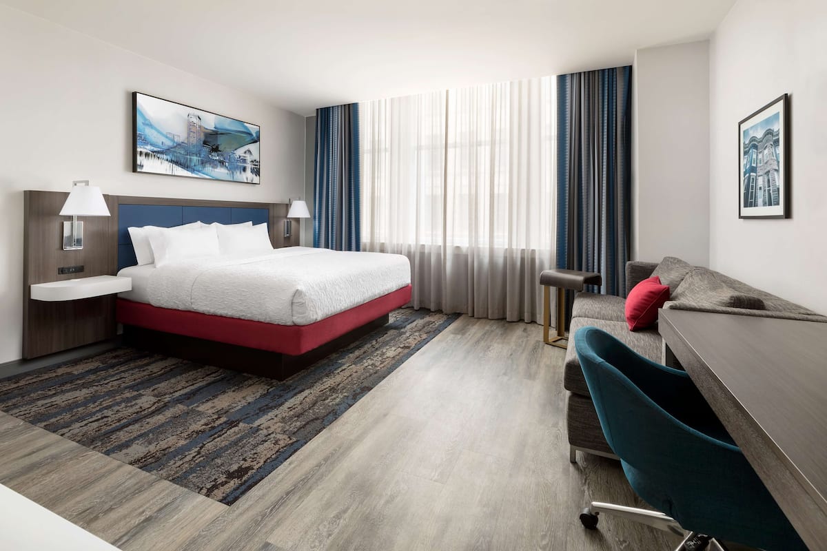Ein Hotelzimmer mit Bett, Schreibtisch, Stuhl, Nachttisch und einem großen Fenster mit Vorhängen.