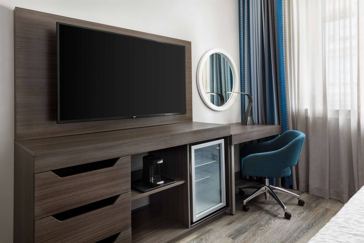 Ein Hotelzimmer mit Flachbildfernseher an einem Holztisch, einer Minikühlschrank, einem blauen Bürostuhl und einem Spiegel an der Wand.