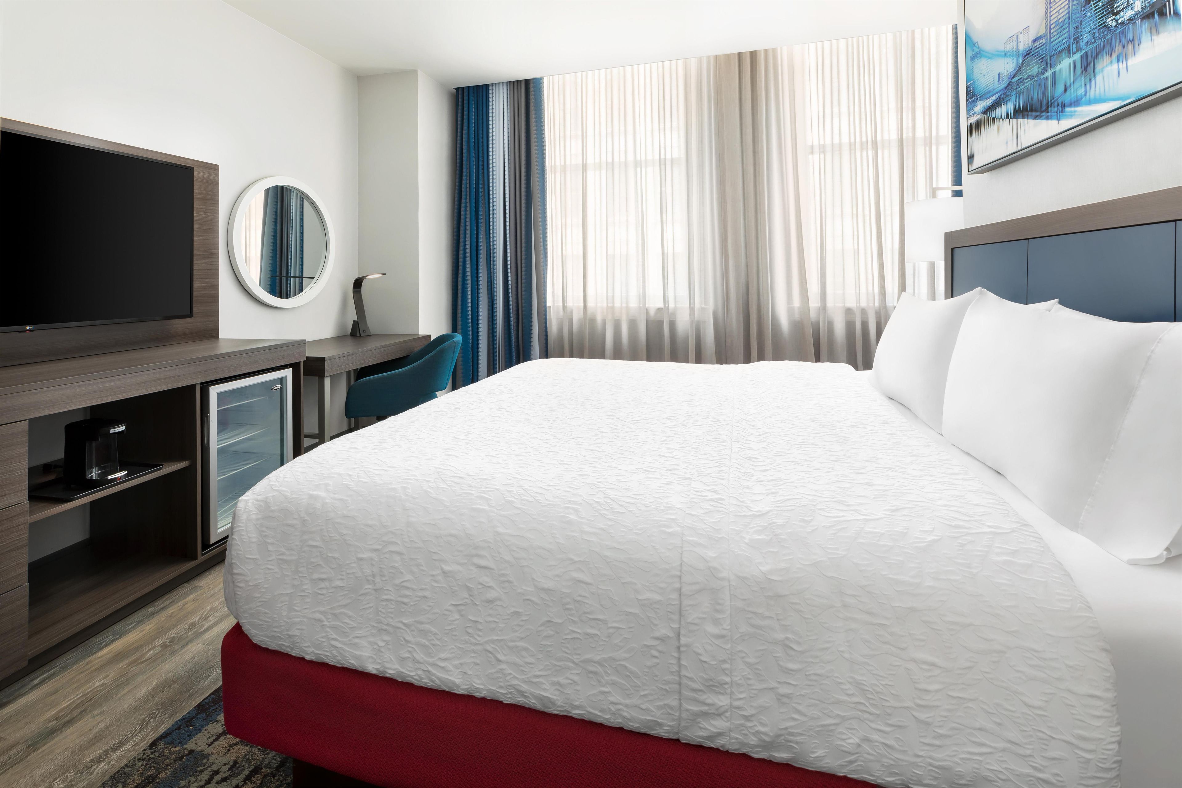 A hotel room with a bed, a TV, a desk with a chair, a coffee machine, and a window with curtains.