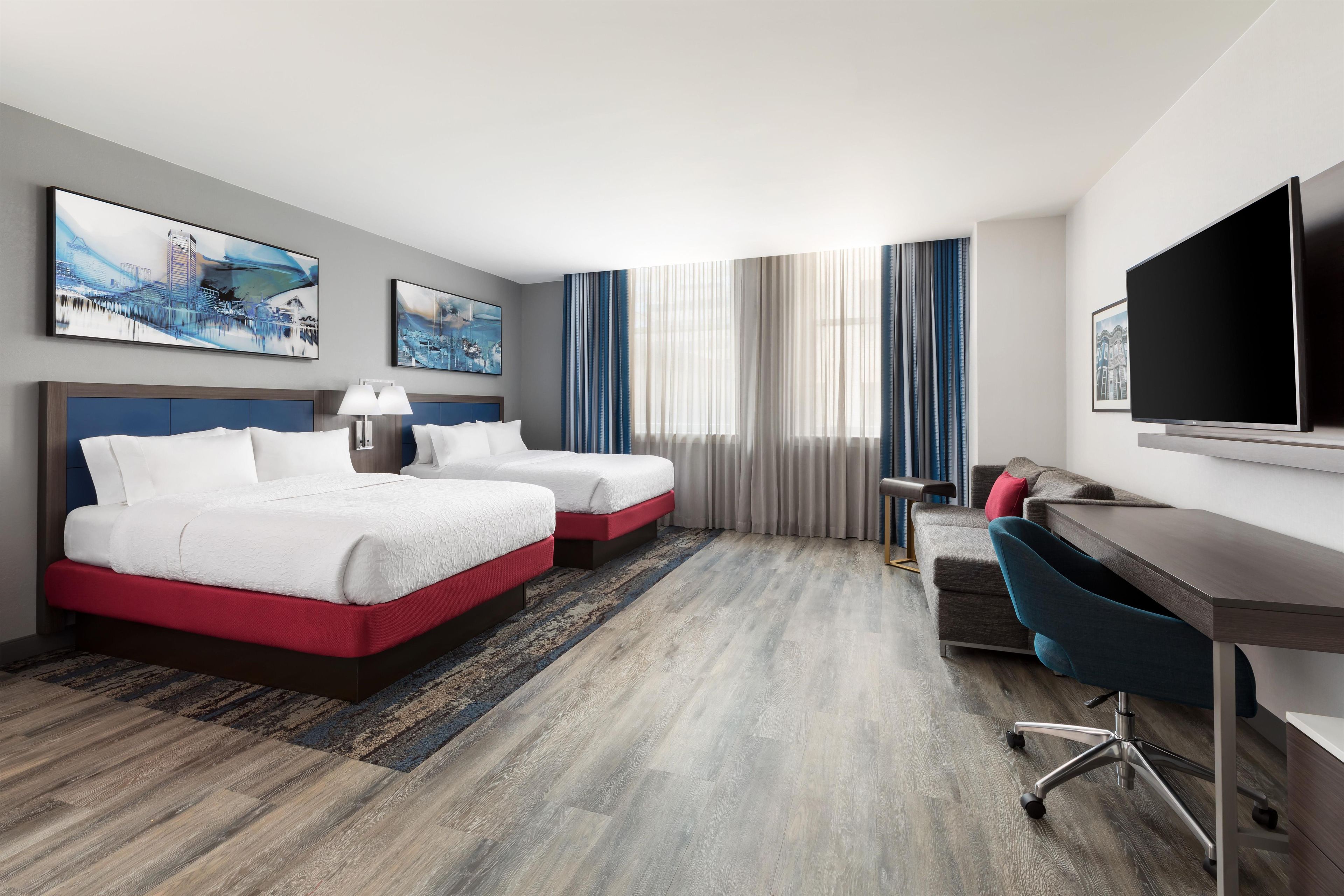 Een hotelkamer met twee bedden, een bureau, een stoel, een televisie en kunstwerken aan de muur.