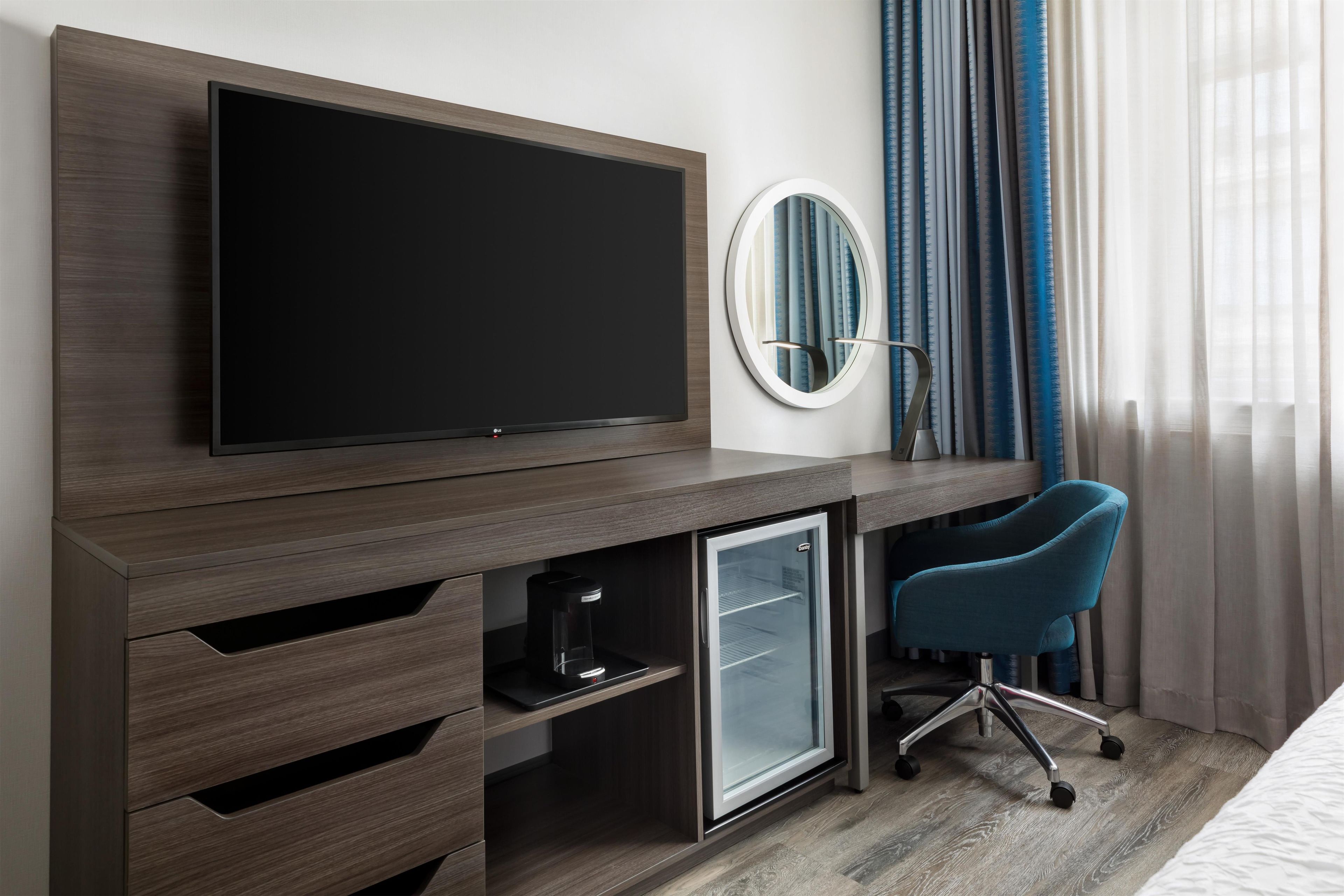 A modern hotel room with a flat-screen TV mounted on a wooden console, a small refrigerator, a desk with a chair, and a mirror.