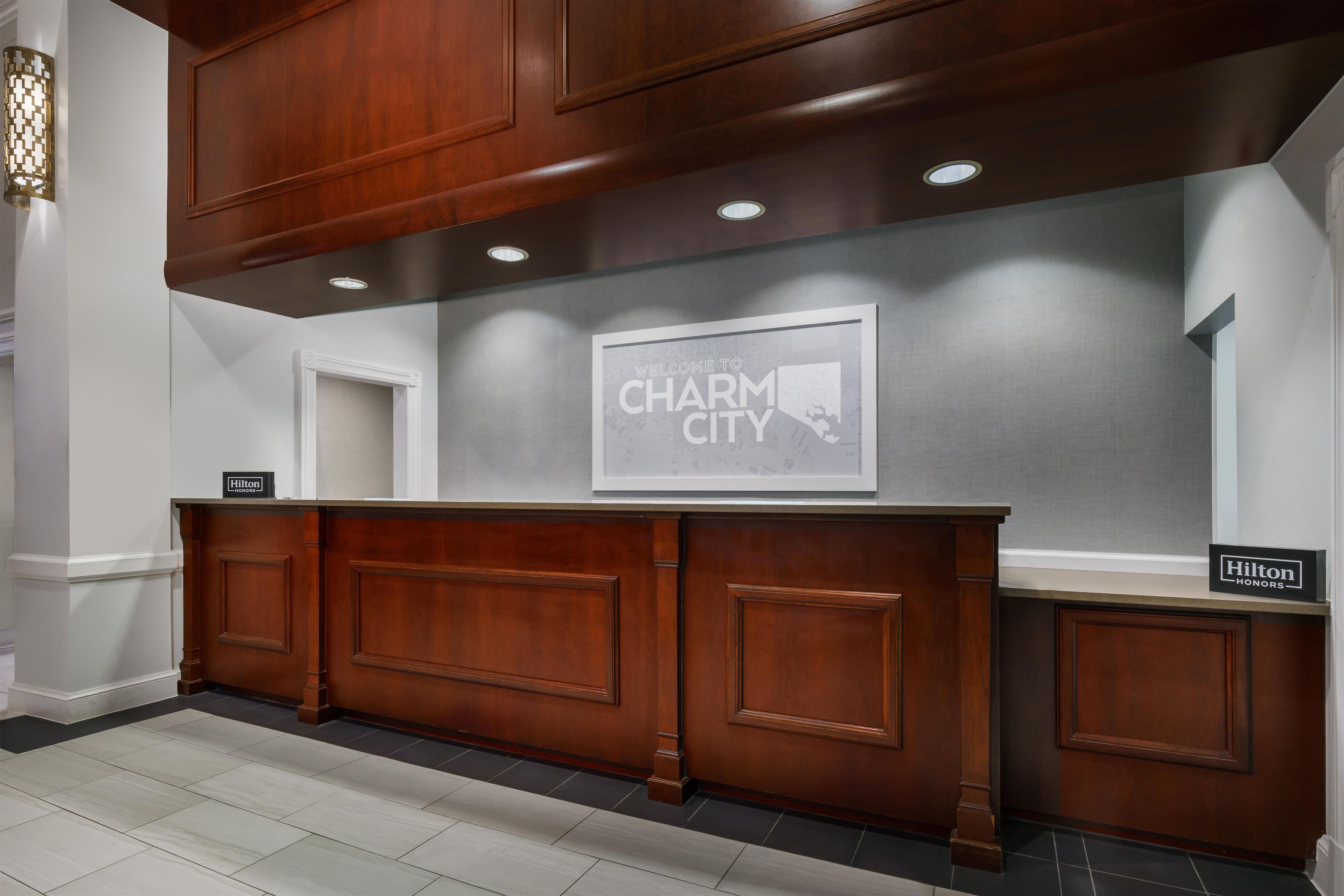 Un mostrador de recepción con un cartel que dice 'Welcome to Charm City', una placa de 'Hilton Honors' y un mapa enmarcado en la pared.
