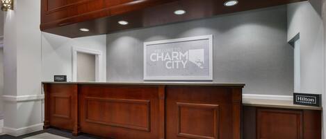 Un mostrador de recepción con un cartel que dice 'Welcome to Charm City', una placa de 'Hilton Honors' y un mapa enmarcado en la pared.