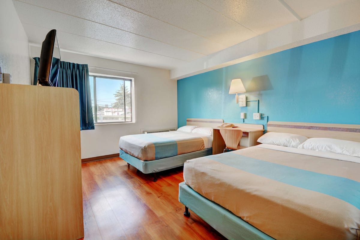 A hotel room with two beds, wooden flooring, a desk, a lamp, and a window with curtains.