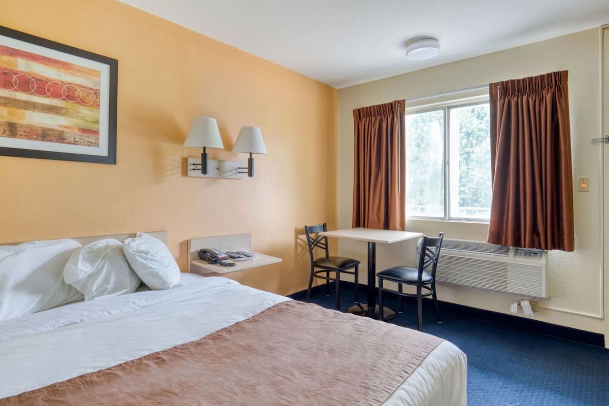 A hotel room with a bed, a small table with chairs, a wall-mounted lamp, a window with curtains, and a radiator.