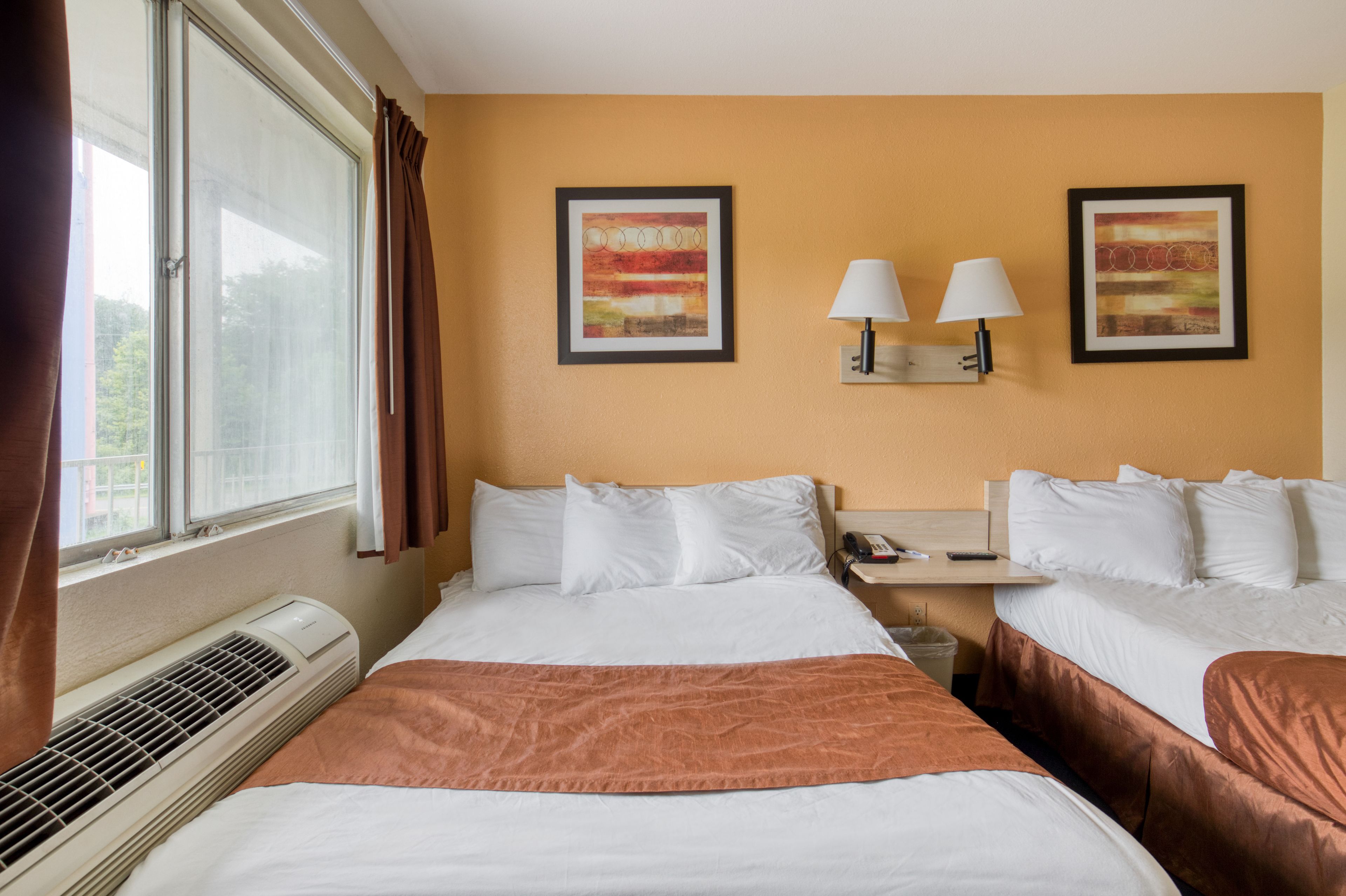 A hotel room with two beds, a window with curtains, an air conditioner, and two wall-mounted lamps.