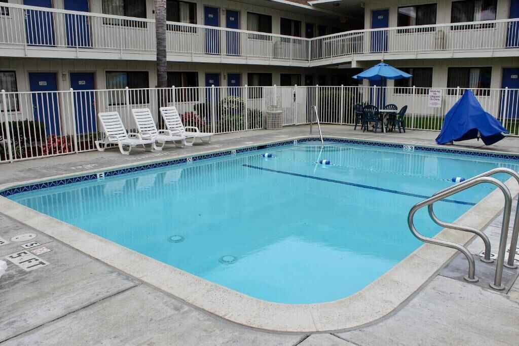 A swimming pool with lounge chairs and a covered seating area, surrounded by a white fence and a multi-story building with balconies.