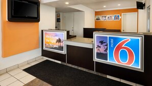 Ein Hotel-Rezeptionsbereich mit modernem Design, der einen Empfangstresen, einen an der Wand montierten Flachbildfernseher und eine digitale Anzeige mit dem Hotelnamen und einer Willkommensbotschaft zeigt.