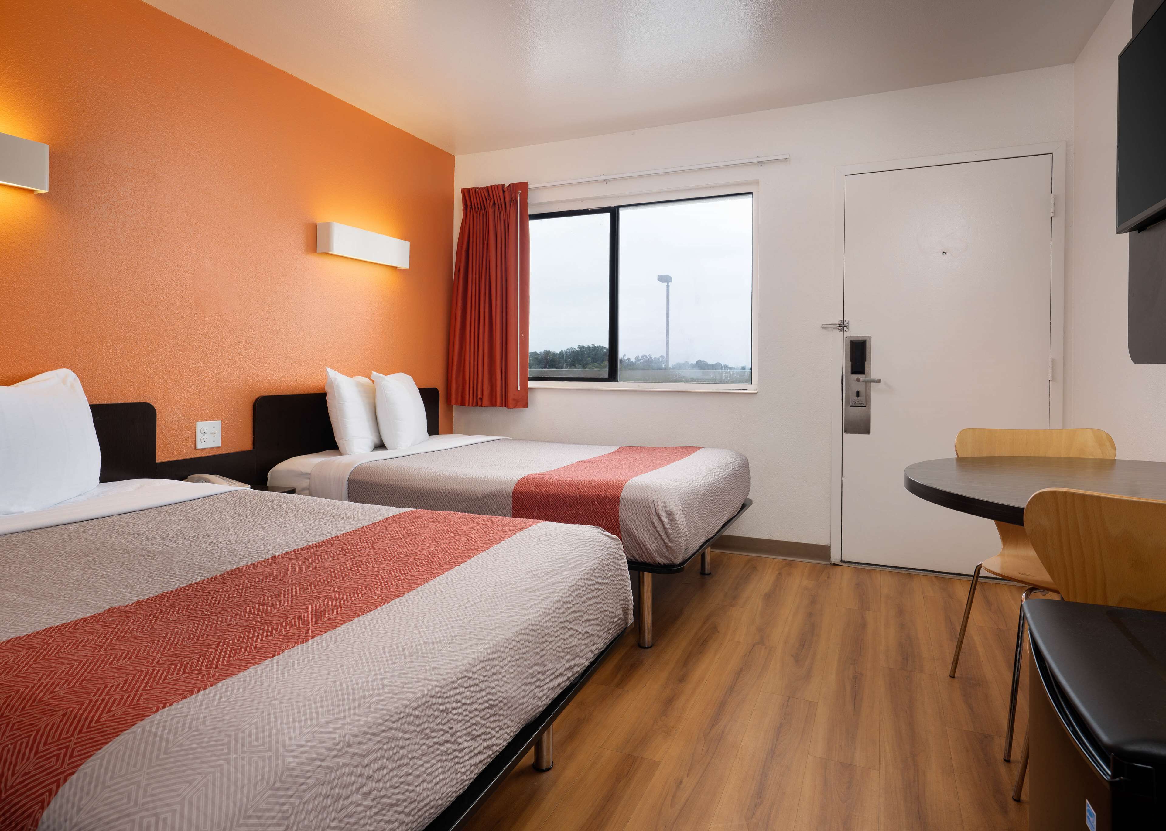 A hotel room with two beds, a small table, a chair, a window with curtains, and a door.