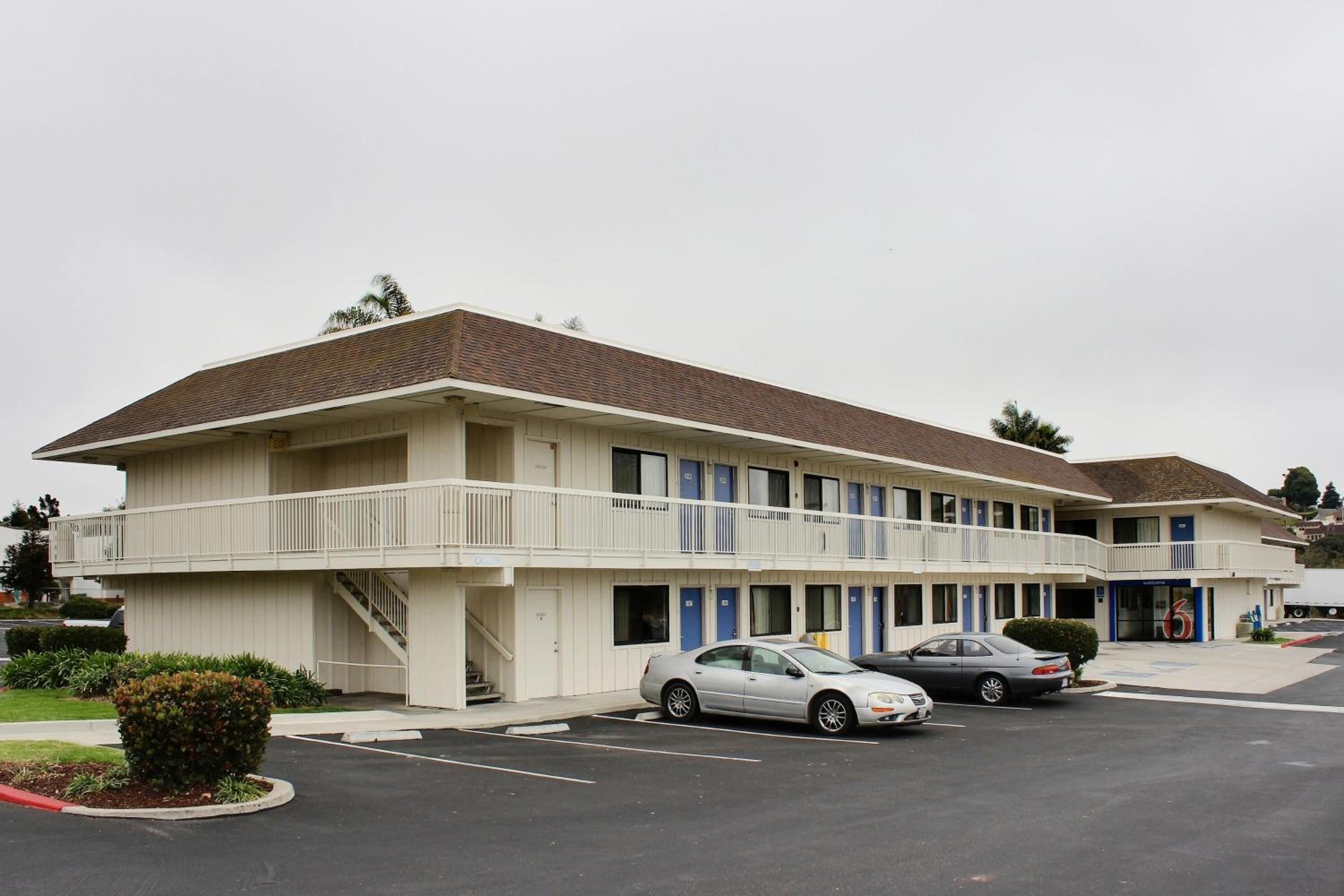 A motel with a two-story structure, a covered parking area, and a small landscaped area with a bush.