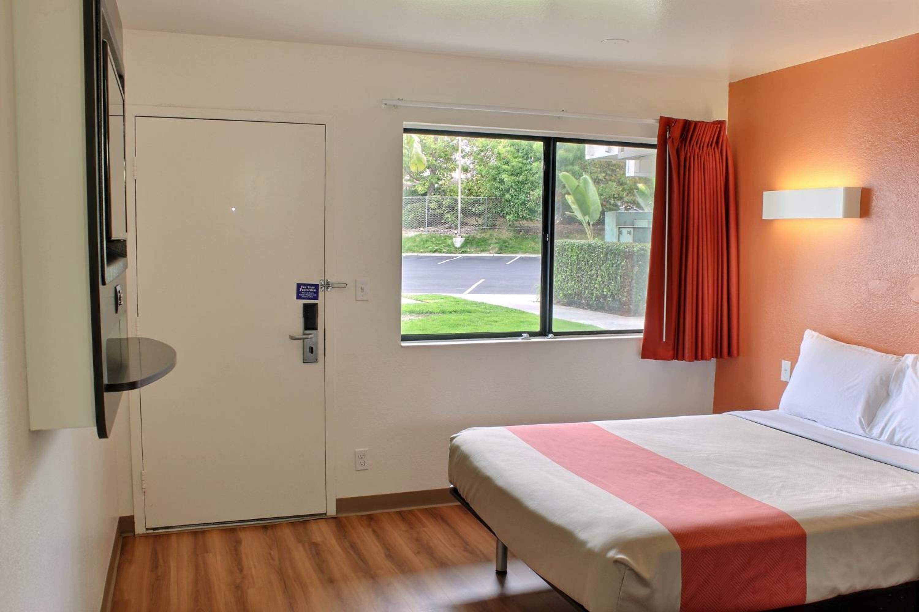 A hotel room with a bed, a window with a view of a parking lot, and a door.