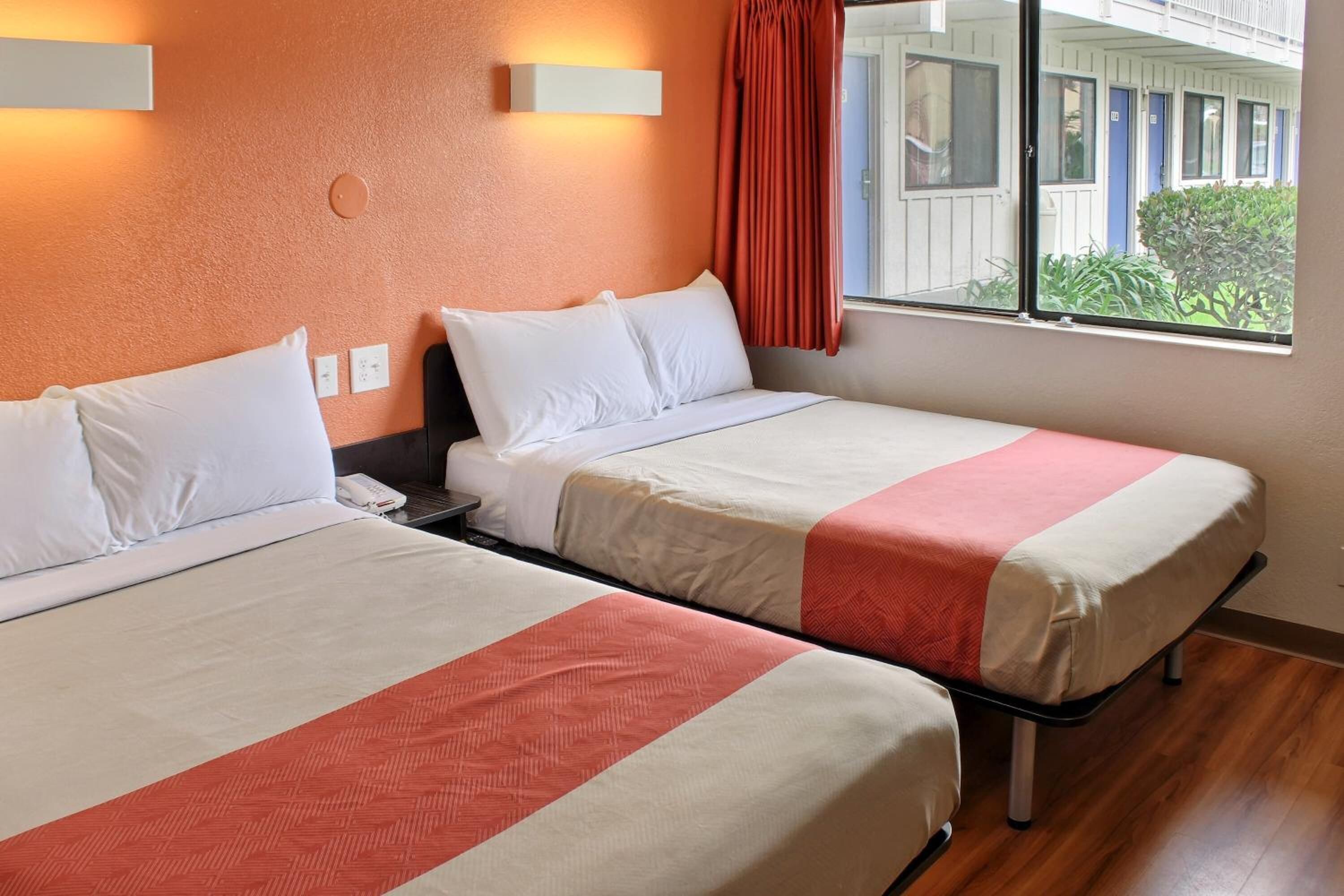 Two single beds with white and red bedding, a bedside phone, and a window with a view of a building and greenery.