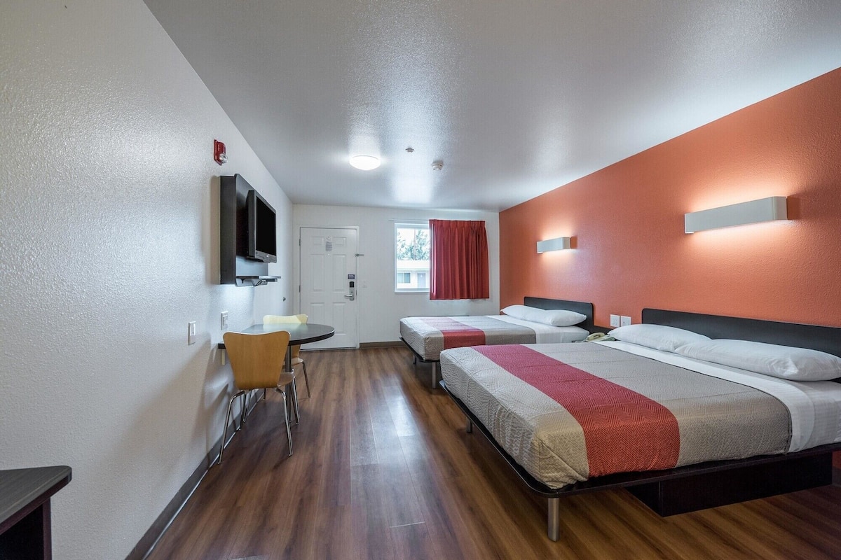 Habitación de hotel con dos camas individuales, una mesa pequeña con una silla, un televisor montado en la pared y una puerta con ventanal.