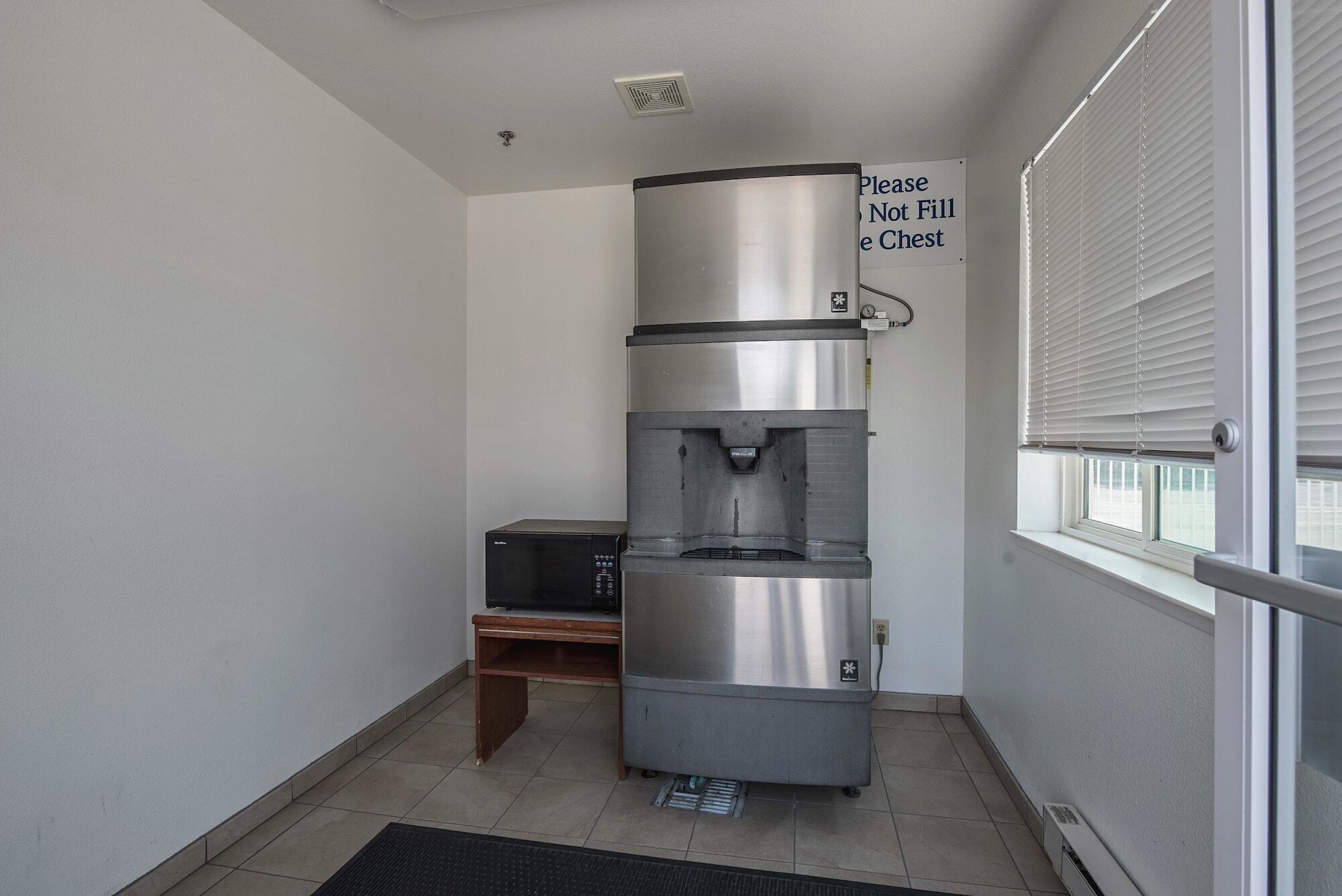 A compact kitchenette with a stainless steel coffee maker, a small wooden table, and a window with blinds.