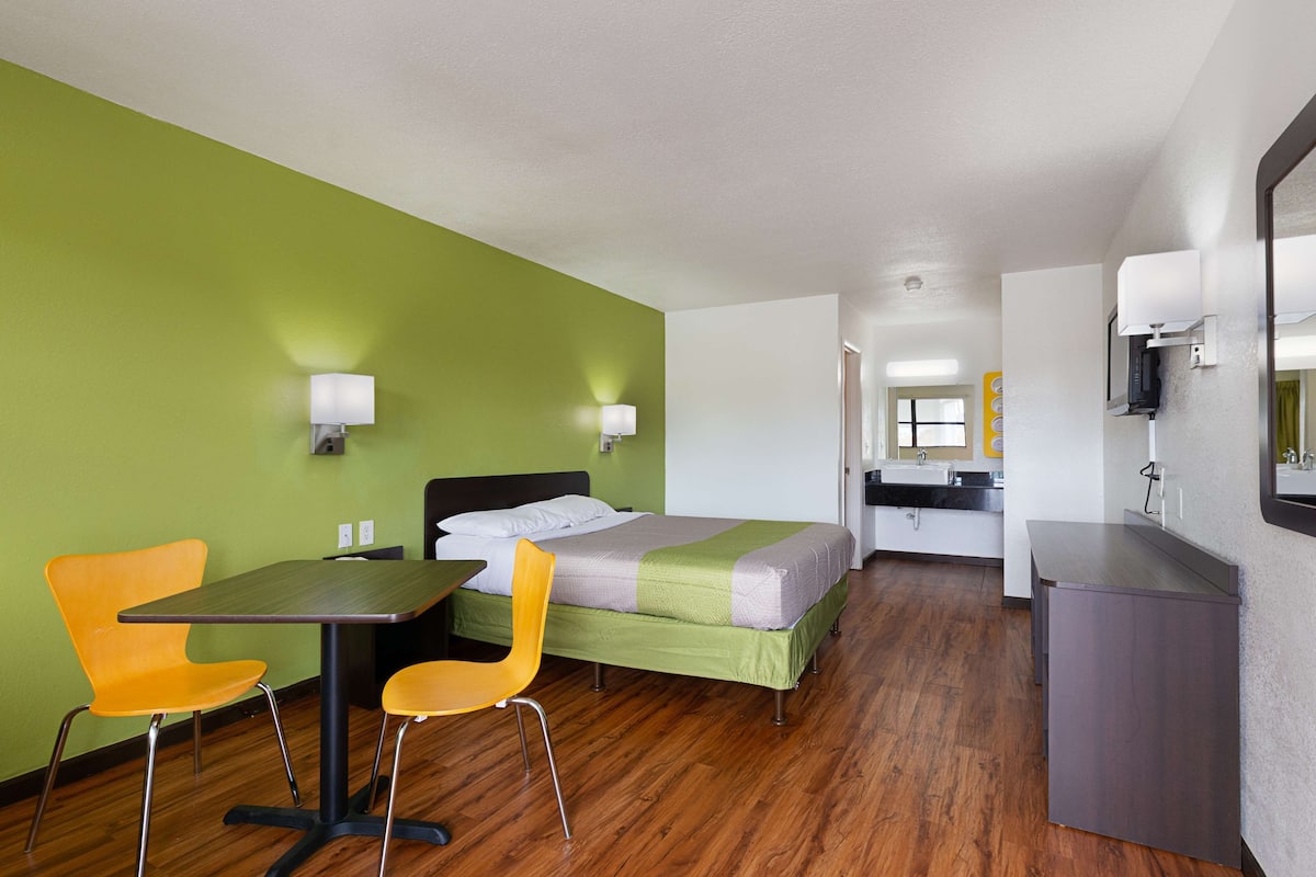 A hotel room with a bed, a desk, two chairs, a small kitchenette, and a bathroom visible in the background.