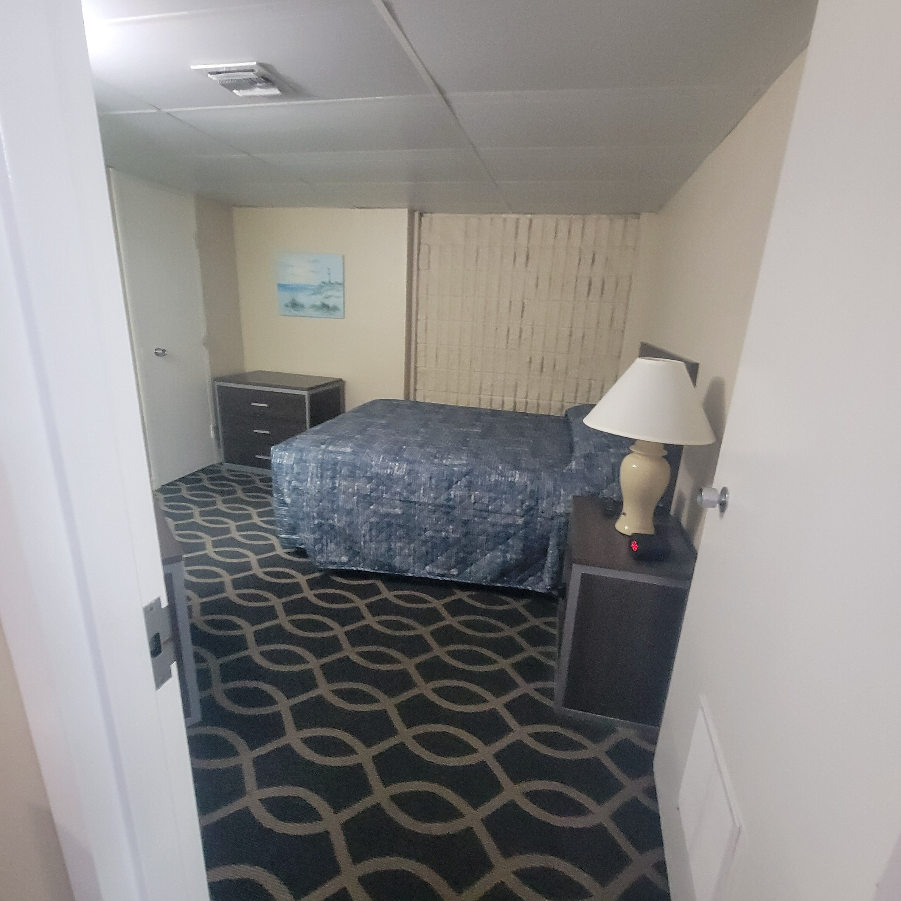 Une chambre d’hôtel avec un lit, une table de chevet avec une lampe, une commode et un tapis à motifs.
