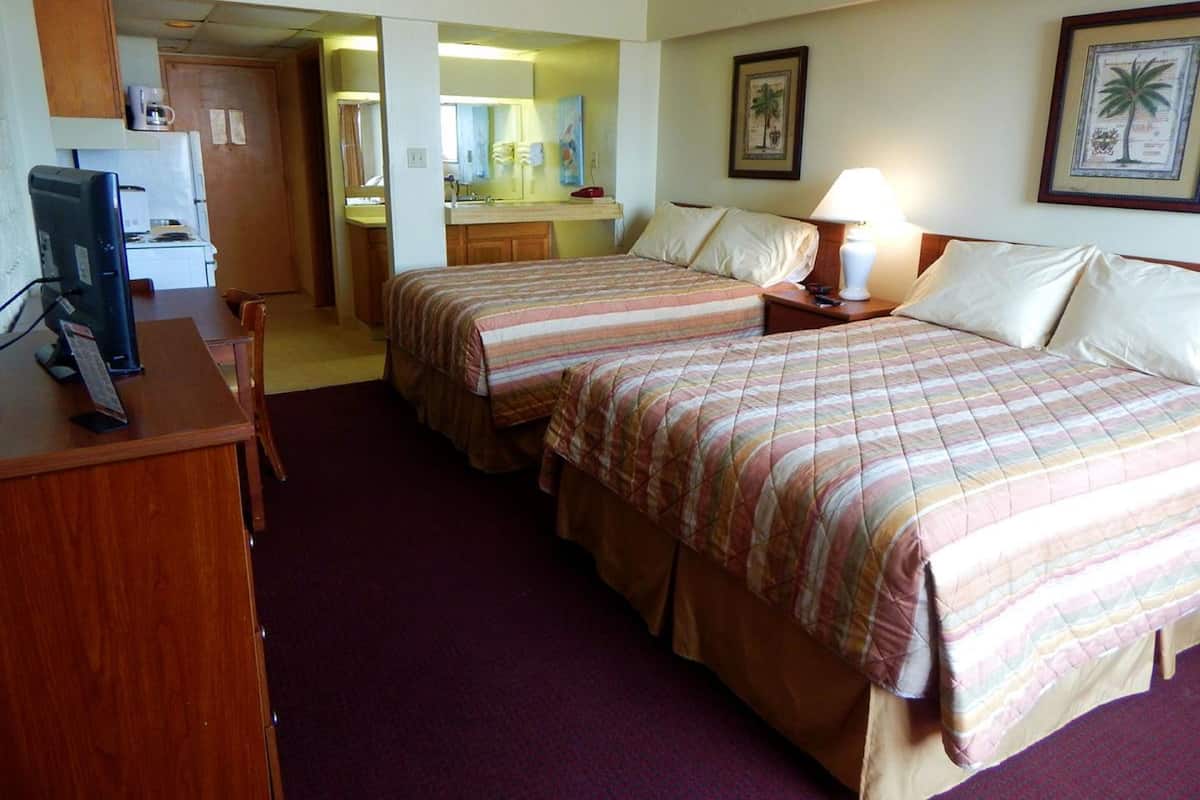A hotel room with two beds, a desk with a computer, a small kitchenette, and a bathroom visible through an open door.