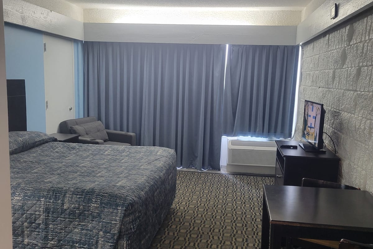 A hotel room with a large bed, a desk, a chair, a TV, and a window with curtains.