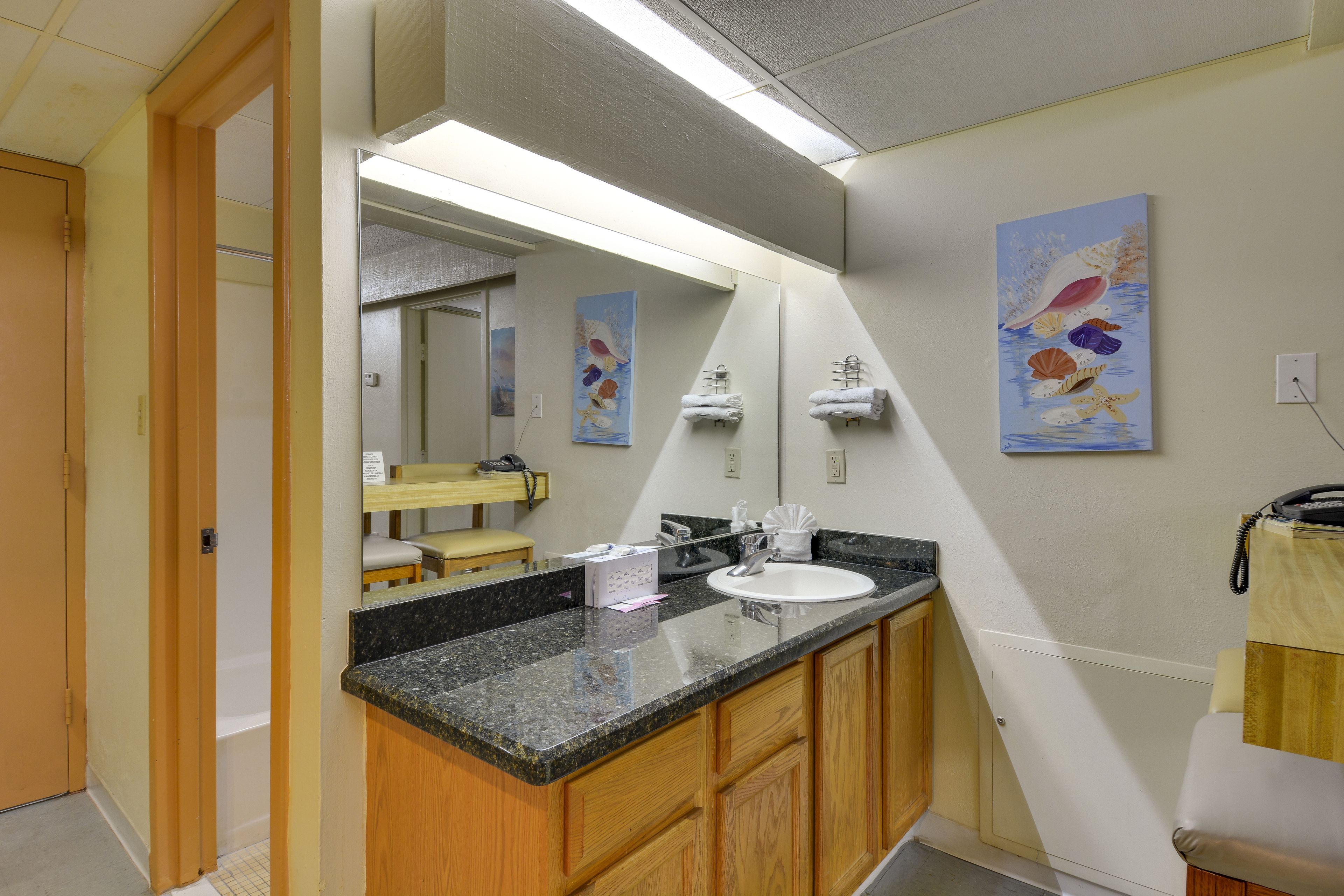 A bathroom with a granite countertop, wooden cabinets, a white sink, and a large mirror. There are two towels hanging on towel racks, and a painting of fish on the wall.