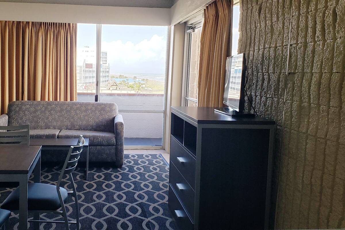 A hotel room with a sofa, dining table, chairs, a TV, and a view of the ocean.