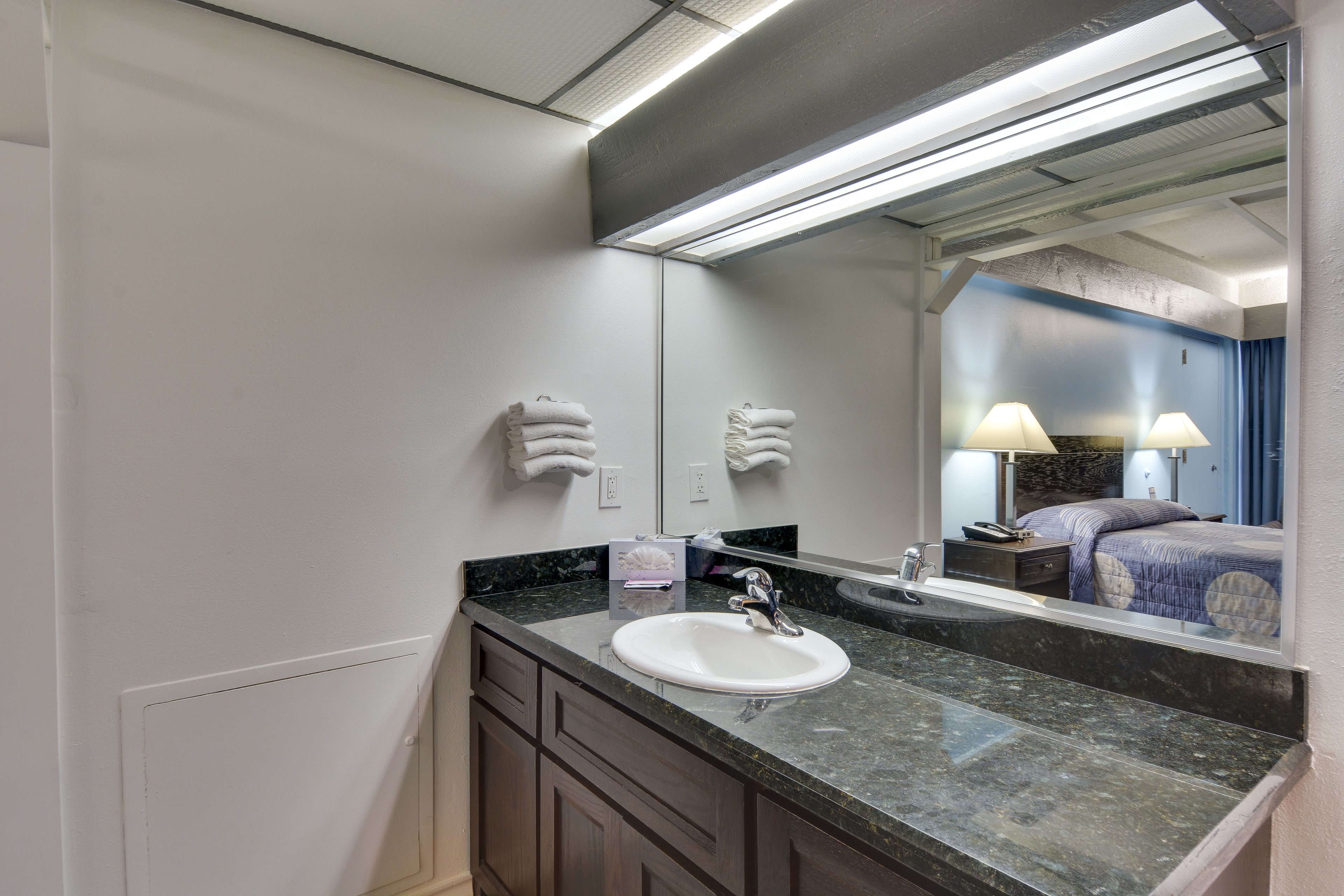A bathroom with a white sink, dark marble countertop, and a large mirror reflecting a bedroom with a bed and nightstand.